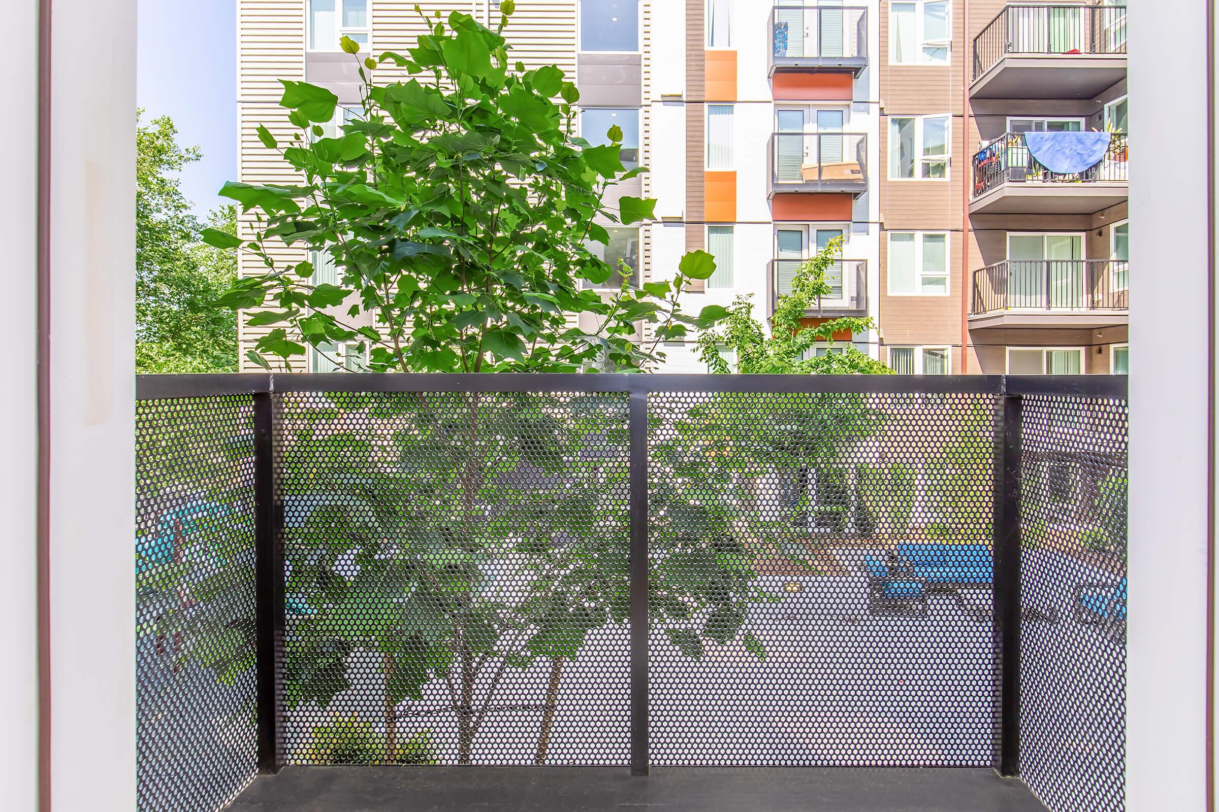View from a balcony featuring green trees and plants. In the background, there are modern residential buildings with balconies. Some outdoor furniture is visible in the courtyard below. The overall atmosphere is open and inviting.