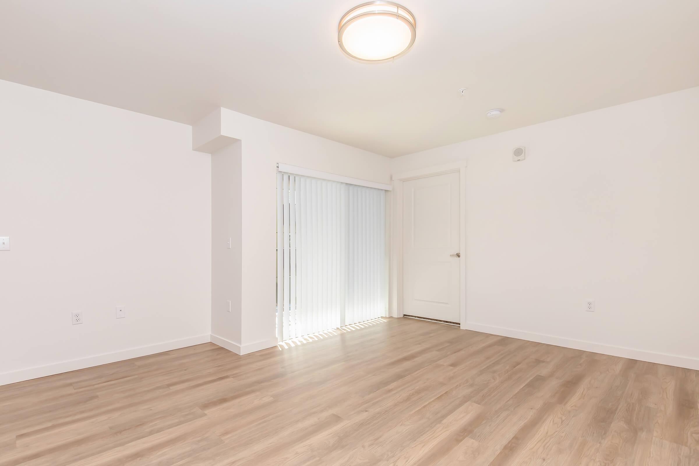 A spacious, empty room with light-colored walls and wooden flooring. A large window with vertical blinds allows natural light to enter, and a modern ceiling light fixture is visible. The room features a door leading outside and a subtle corner where the wall extends.