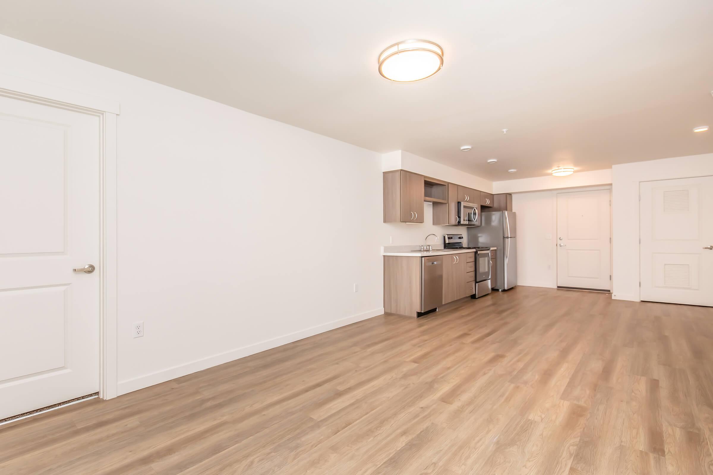 Interior view of a modern apartment showcasing a spacious, open layout. The image features light wood flooring, a kitchen area with stainless steel appliances, and white cabinetry, along with a bright, neutral color scheme. A doorway is visible, leading to another room or entrance.