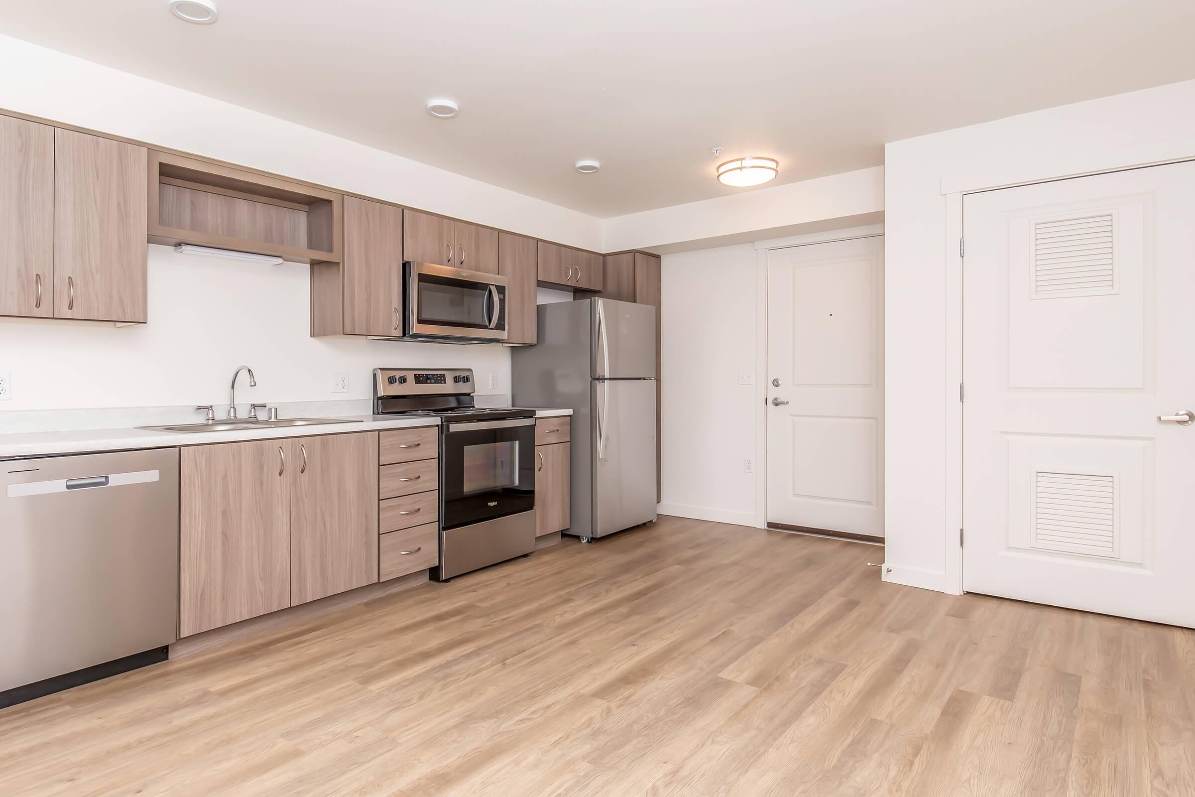 Modern kitchen with wooden cabinets, stainless steel appliances, a microwave, and a dishwasher. The open layout features a countertop and a view of a front door. Light-colored flooring and bright lighting create a spacious, inviting atmosphere.