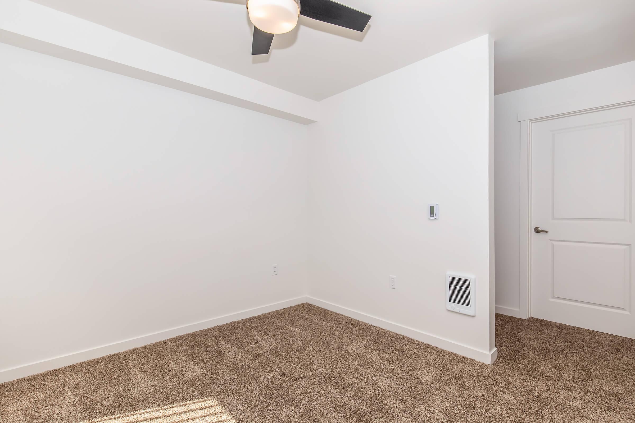 Empty interior room with light beige carpet, white walls, and a ceiling fan. A door is visible on the right side, and a wall vent is located near the floor. The room is well-lit with natural light coming from a window, creating a neutral and clean aesthetic.
