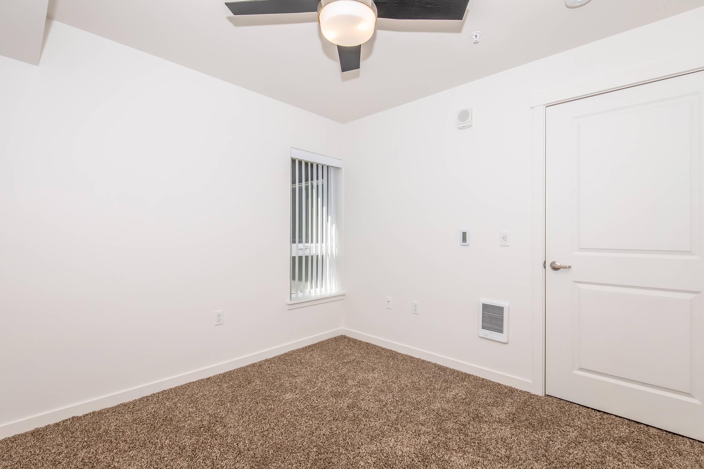A spacious, empty room with light beige carpet, a ceiling fan, and a window with vertical blinds. The walls are painted white, and there is a door leading to another area. An air vent is visible on one wall, and the room is well-lit and airy.