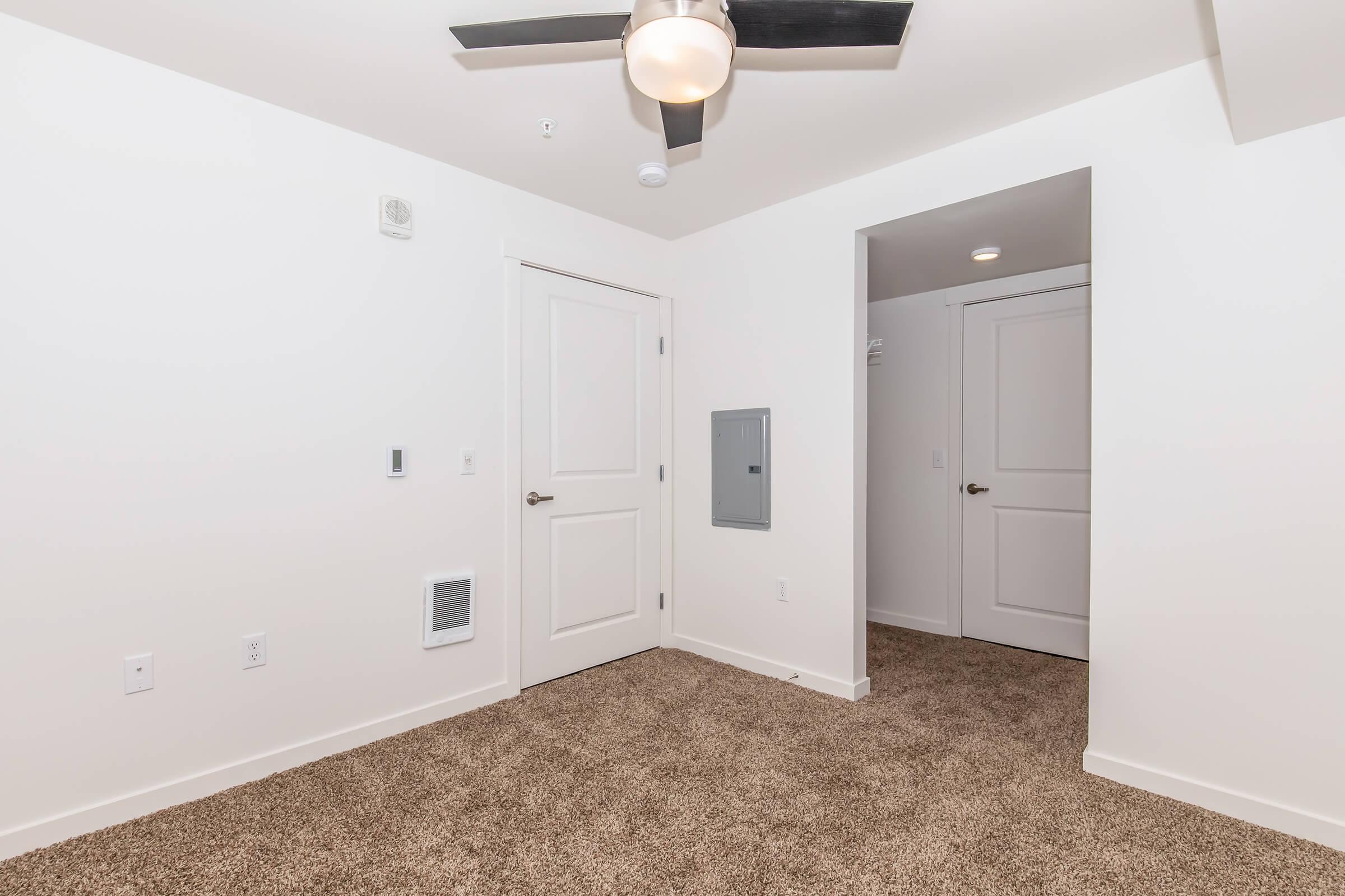 A spacious, empty room with light beige carpet. The walls are painted white, and there is a ceiling fan above. A door leads to another room on the right, and a utility panel is mounted on the wall. The room is well-lit, with no furniture present, creating a clean and minimalistic atmosphere.