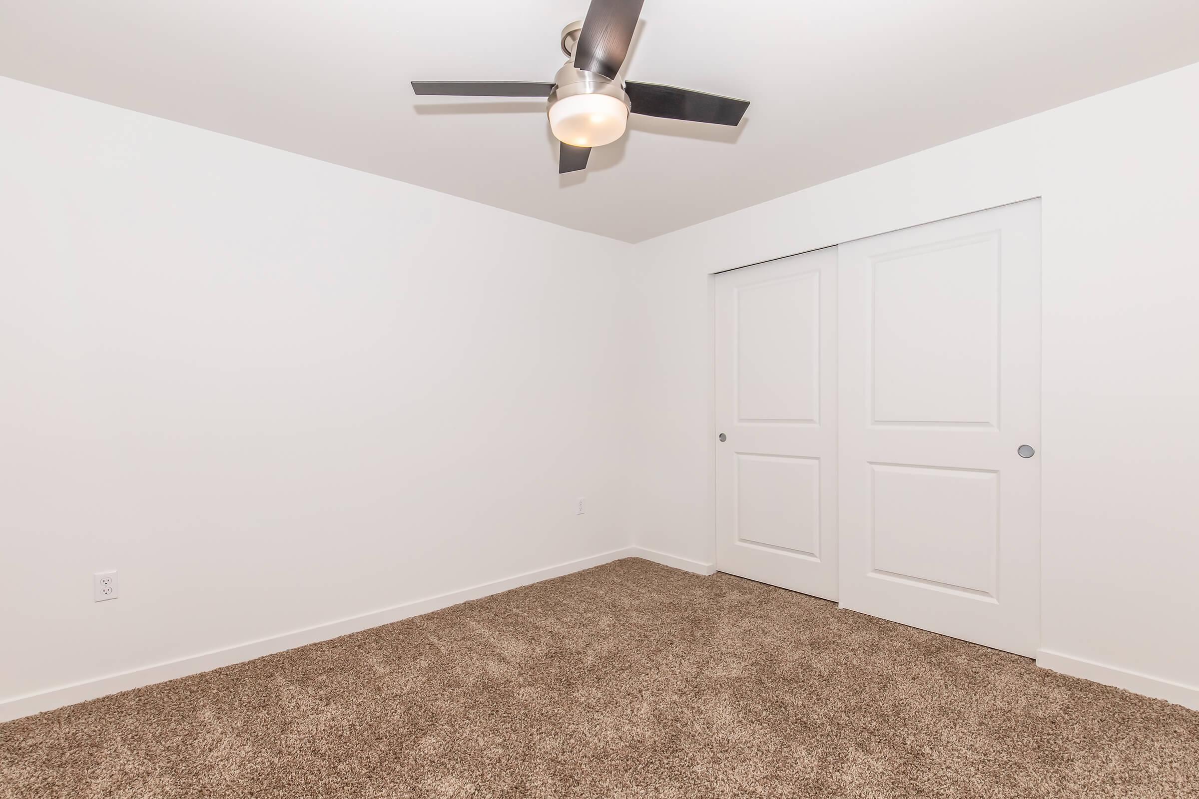 A clean, empty room with light-colored walls and a plush carpet. The room features a modern ceiling fan and a large closet with double doors. The space is bright and minimalist, ideal for various uses such as a bedroom or office.