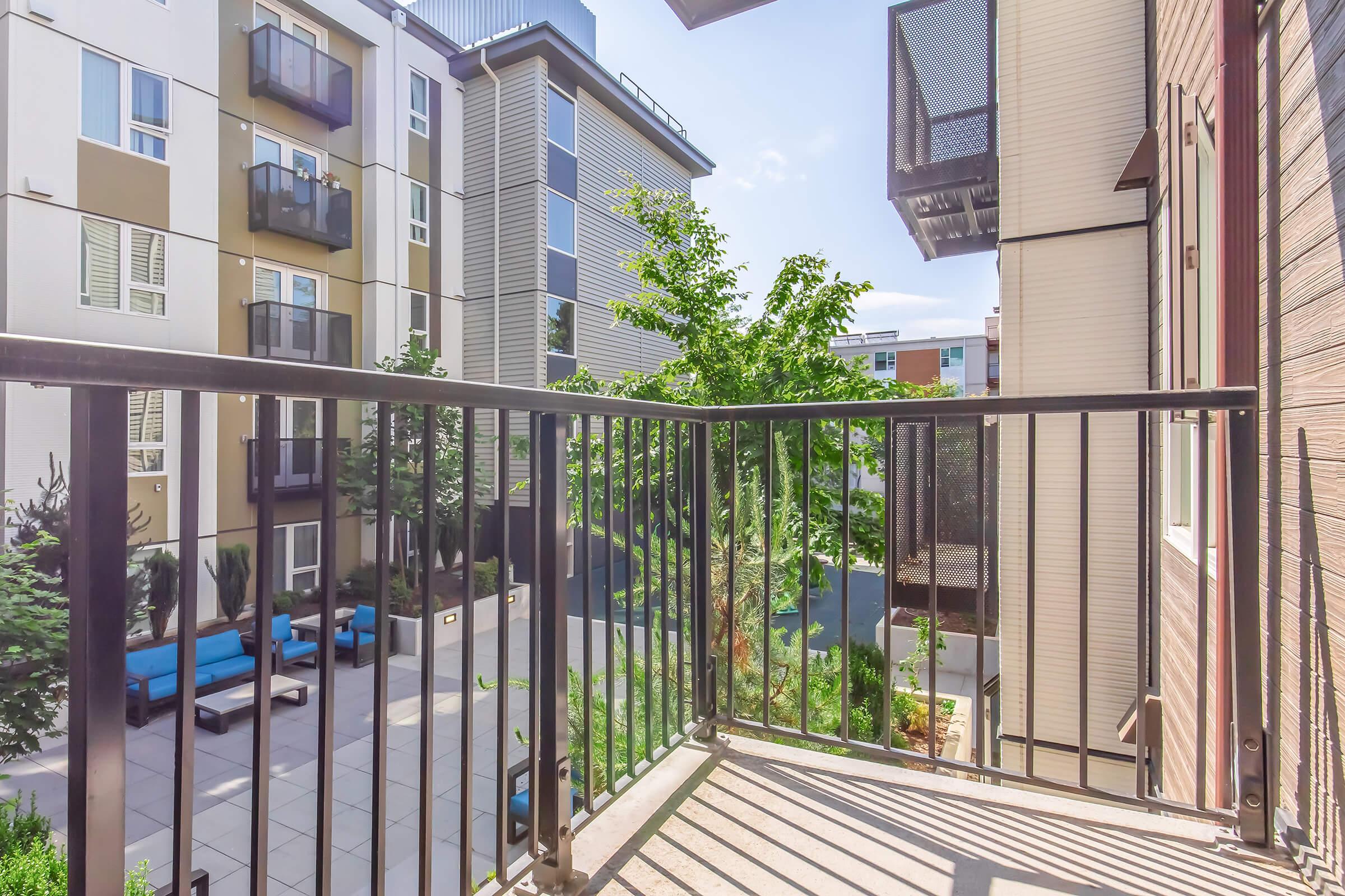 View from a balcony overlooking a landscaped courtyard with green trees and outdoor seating. The scene includes modern apartment buildings in the background and a clear sky, creating a serene urban atmosphere.