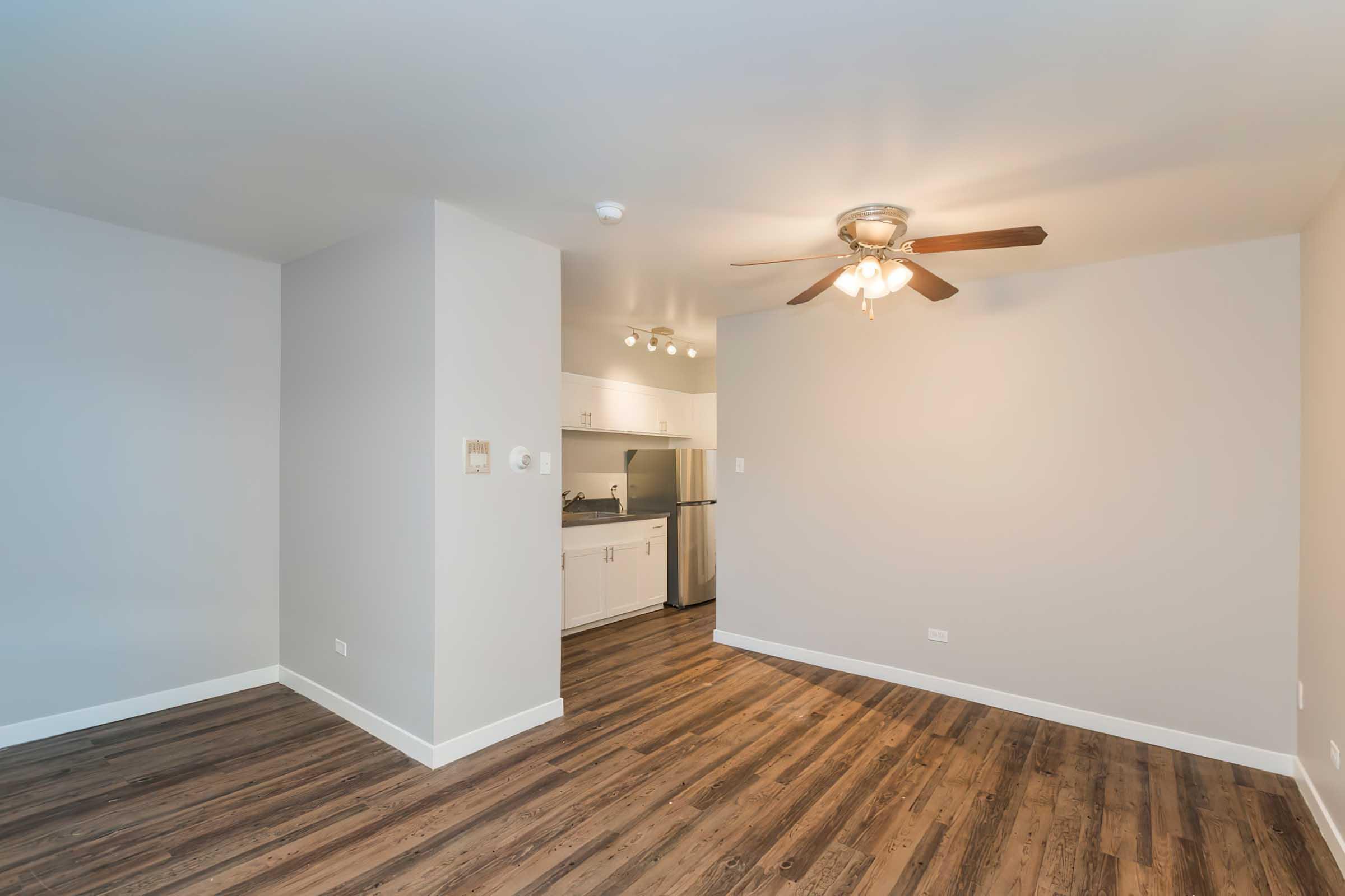 nterior view of a modern apartment living space featuring a ceiling fan, hardwood-style flooring, and a light-colored wall. 