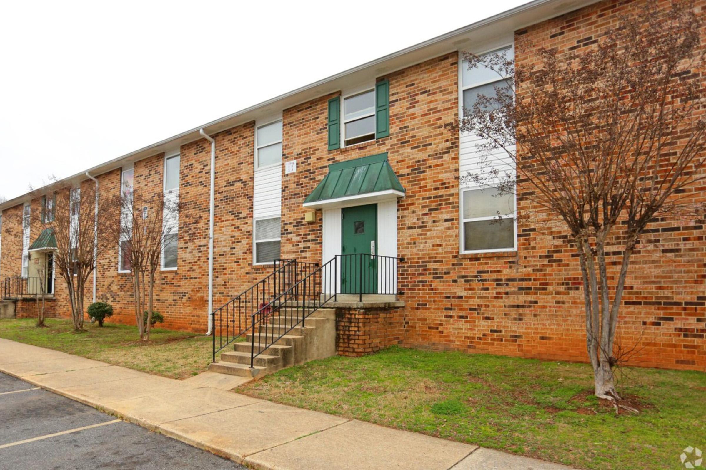 A two-story brick apartment building with green shutters and a green roof over the entrance. The building features multiple windows, a set of stairs leading to the front door, and small shrubs planted near the entrance. The surrounding area has a grassy lawn and a paved parking space.