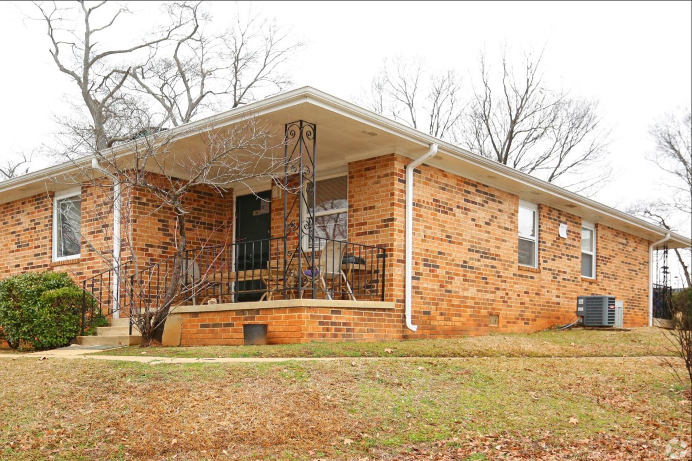 A brick house with a covered porch, featuring black railings and a small shrub in the front yard. The landscape is mostly bare, with a few trees in the background and patches of grass. The house has several windows and an air conditioning unit on the side. The scene is captured on a cloudy day.