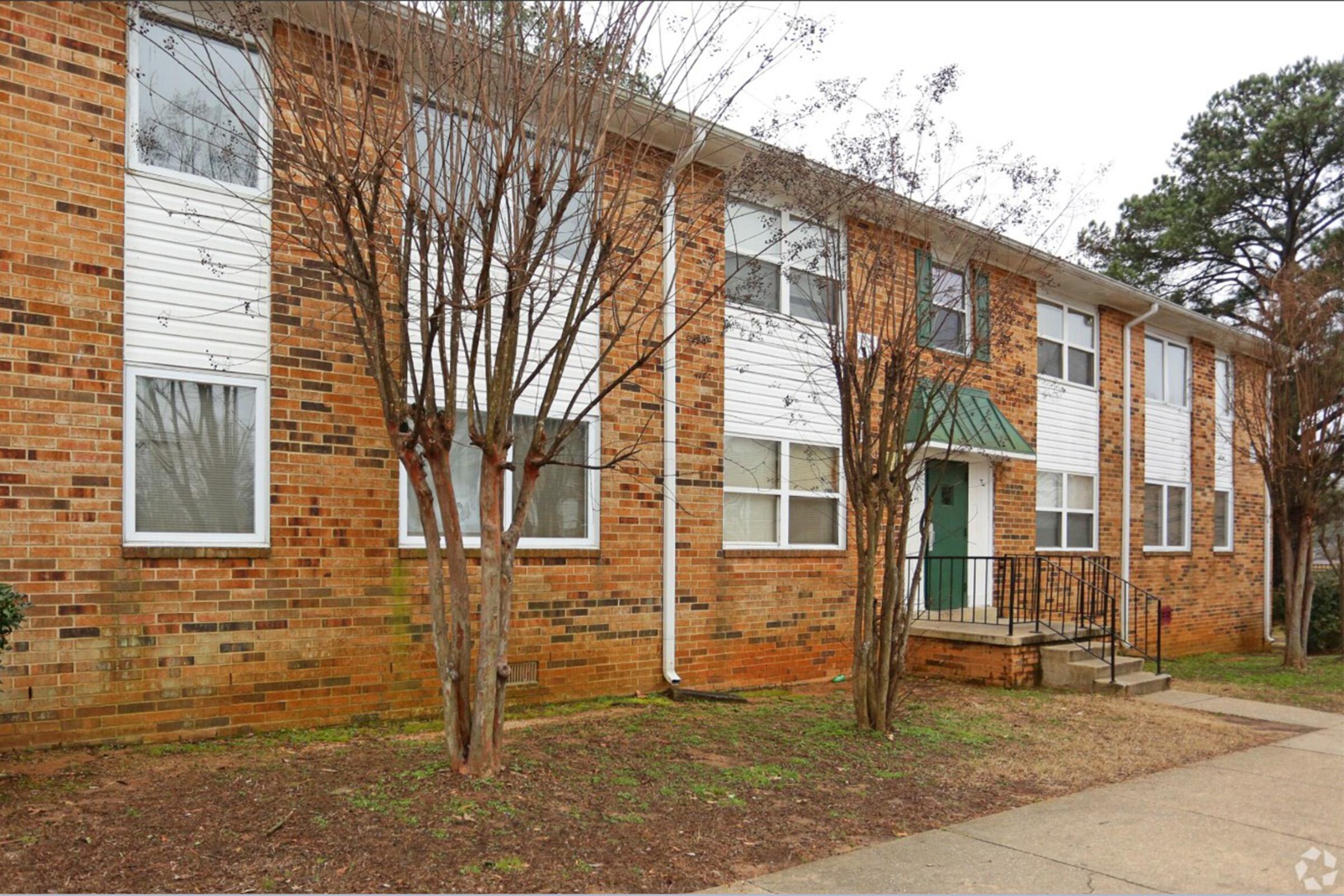 A brick apartment building with white siding accents and multiple windows. The entrance has a green door and a small awning, with a staircase leading up to it. There are small trees in front and a grassy area surrounding the building, set in a residential environment.