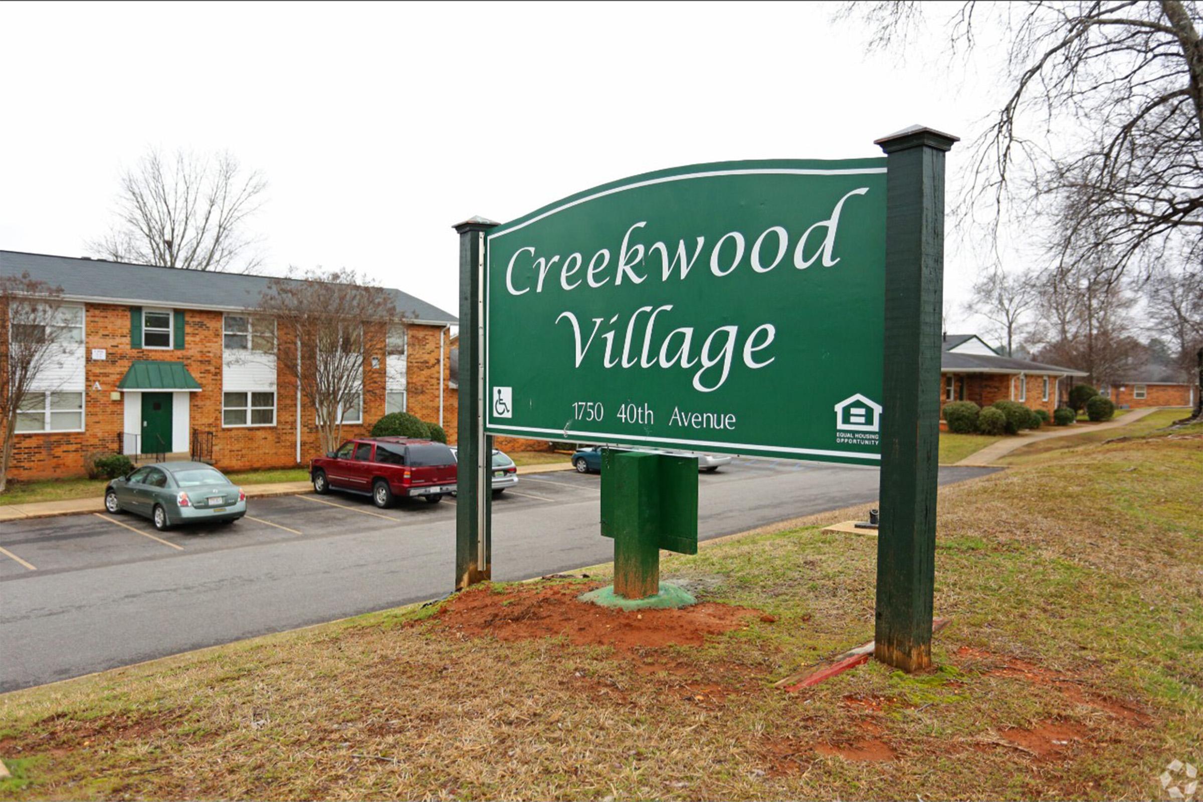 Sign for Creekwood Village located on 40th Avenue, featuring a green background with white text. The area includes several residential buildings, some parked cars, and grassy surroundings, indicating a community environment. The image captures a cloudy day.