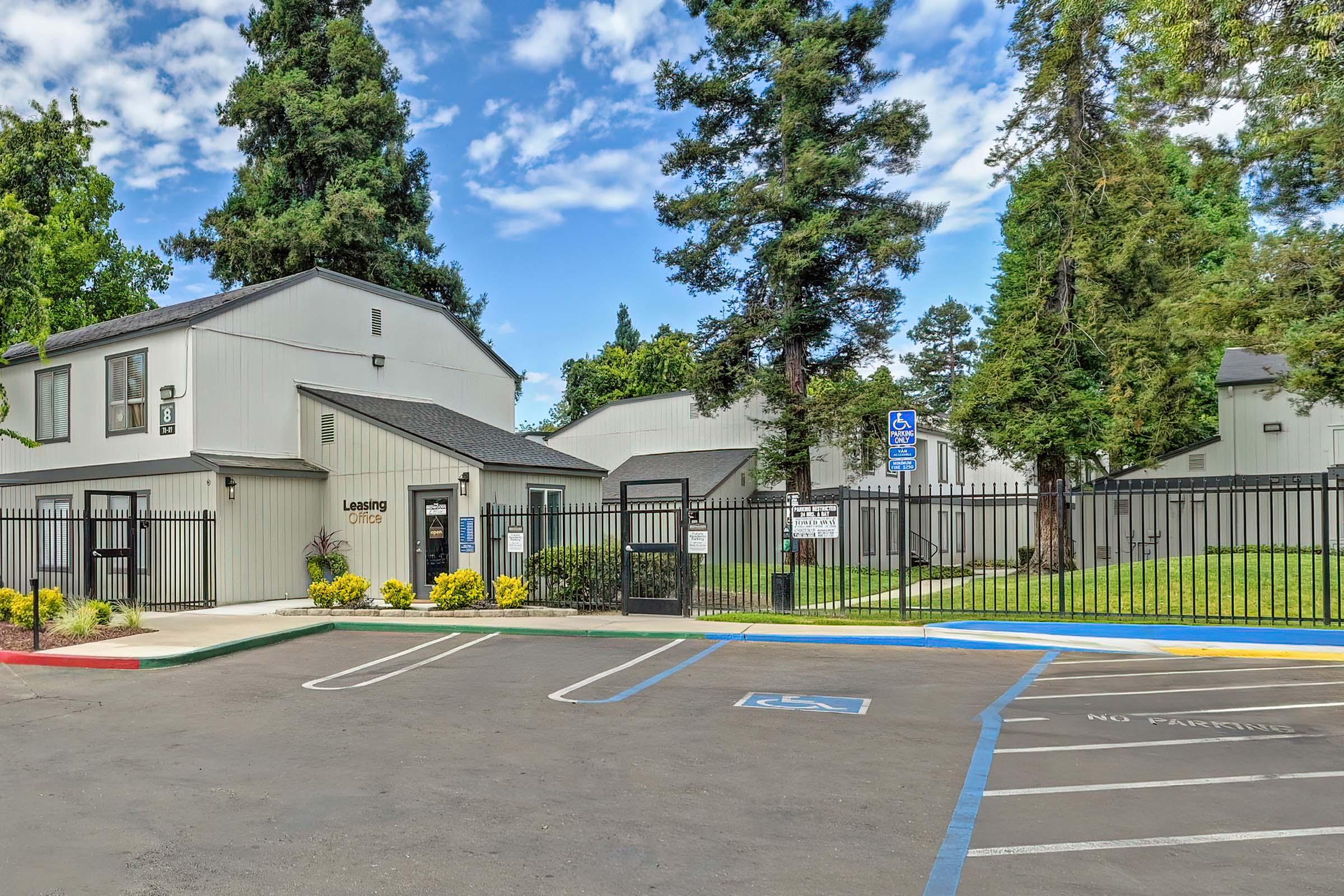 Exterior view of a multi-building residential complex with a leasing office. The area is surrounded by a black metal fence and features well-maintained landscaping, including trees and flowers. The parking lot includes designated spaces, with a sign indicating accessibility. Bright blue sky with scattered clouds in the background.