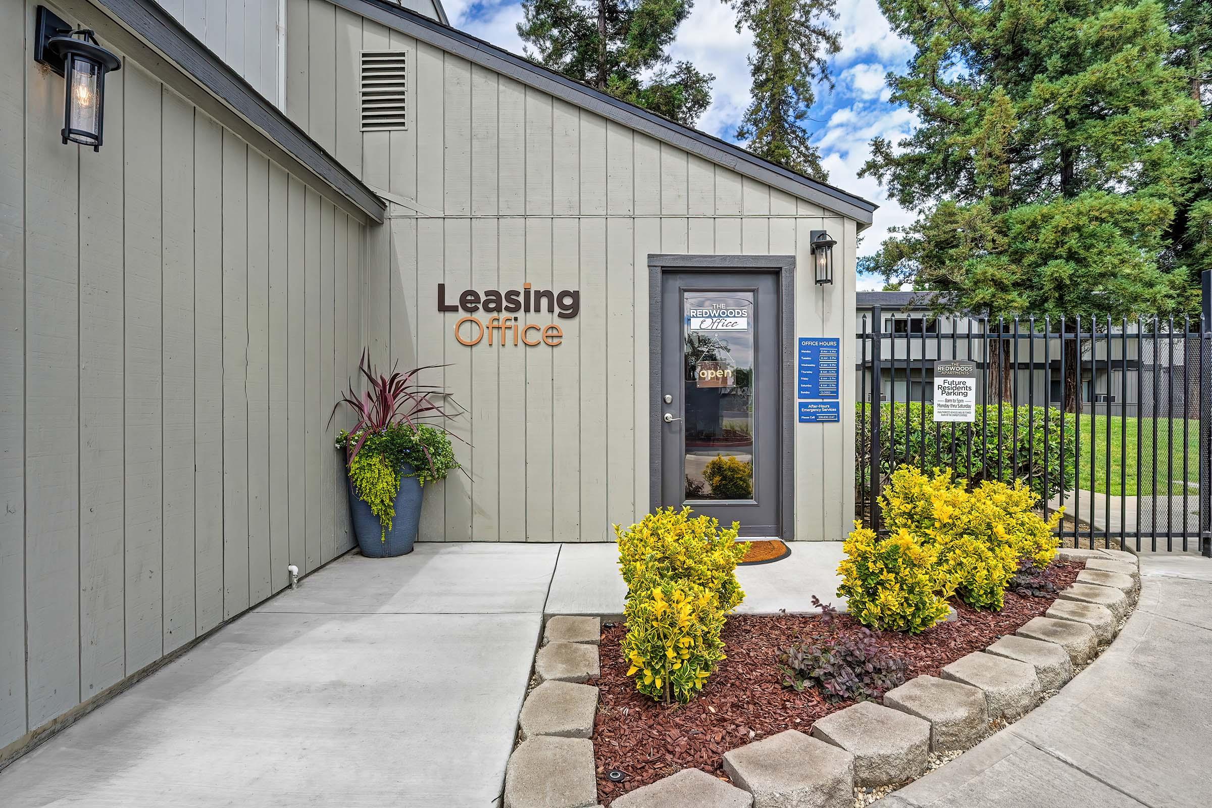 Exterior of a leasing office featuring a gray building with a welcoming entrance. A sign indicates "Leasing Office," and there's a door with a "Open" sign. Surrounding greenery includes potted plants and landscaped bushes. The pathway is lined with stones leading to the entrance, and a fence is visible in the background.