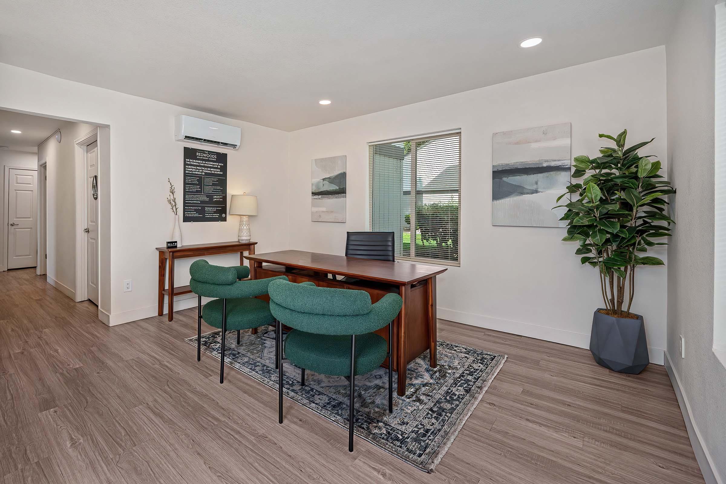 A modern office space featuring a wooden desk with a black chair and two green upholstered chairs. The room has light-colored walls, large windows with blinds, and a potted plant in the corner. Two abstract paintings adorn the wall, and the flooring is a light wood finish.