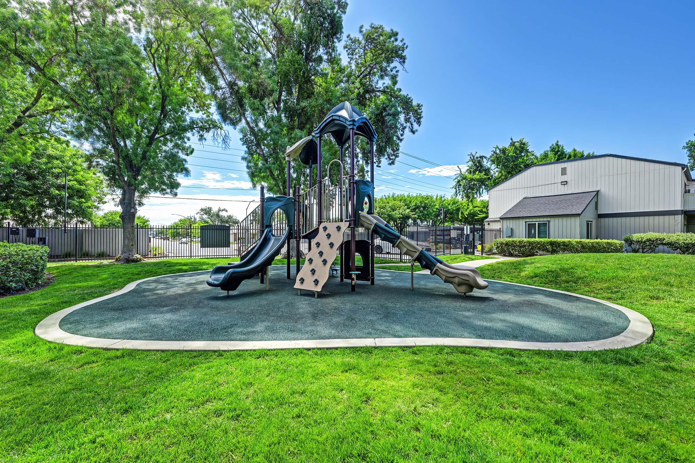 A playground featuring a modern play structure with slides, situated on a grassy area surrounded by trees. The bright blue sky and well-maintained environment create a cheerful atmosphere, ideal for children to play and explore.