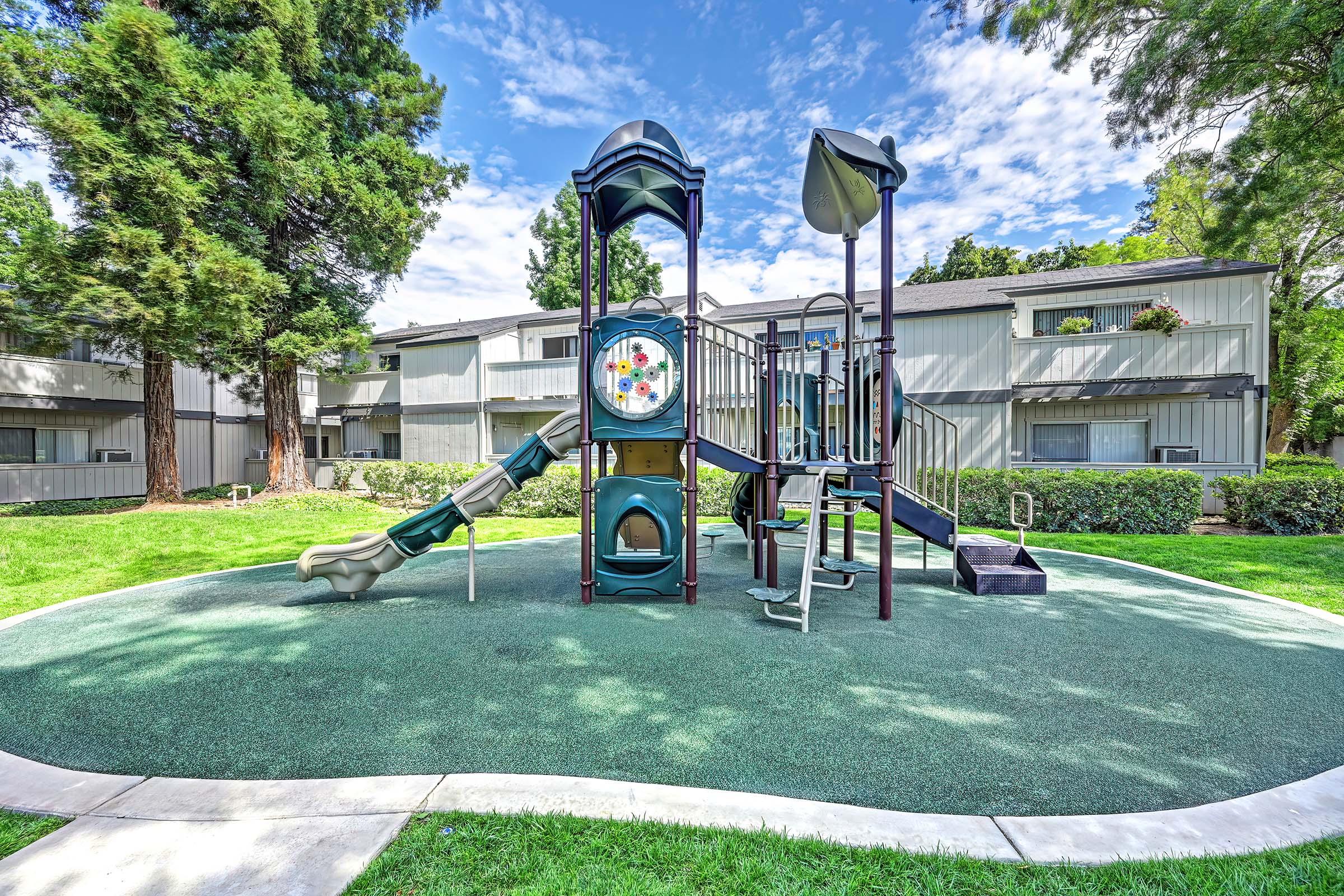 A colorful playground with slides, climbing structures, and a spinning wheel, situated on a grassy area surrounded by trees. In the background, there are residential buildings with balconies. The sky is mostly clear with some clouds.