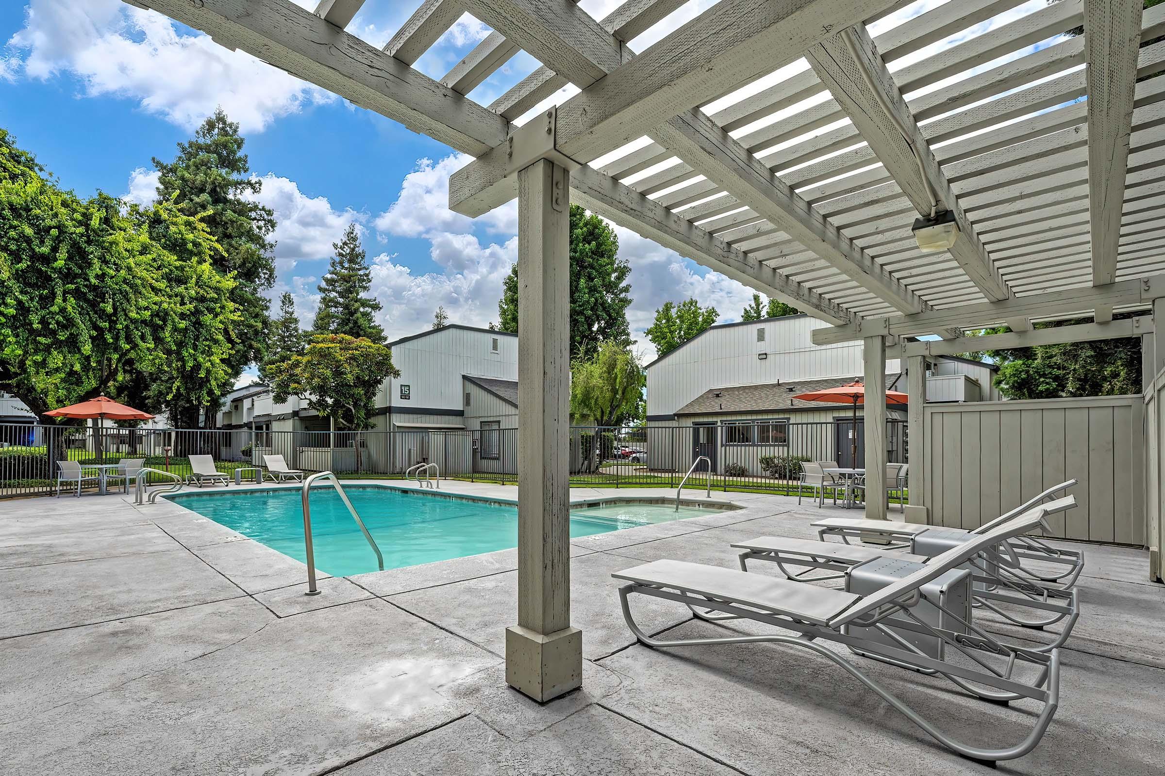 A serene swimming pool surrounded by lounge chairs and shaded by a pergola, with green trees in the background. The area is bright and inviting, featuring blue skies with scattered clouds and orange umbrellas providing additional shade. The pool area is well-maintained and perfect for relaxation.
