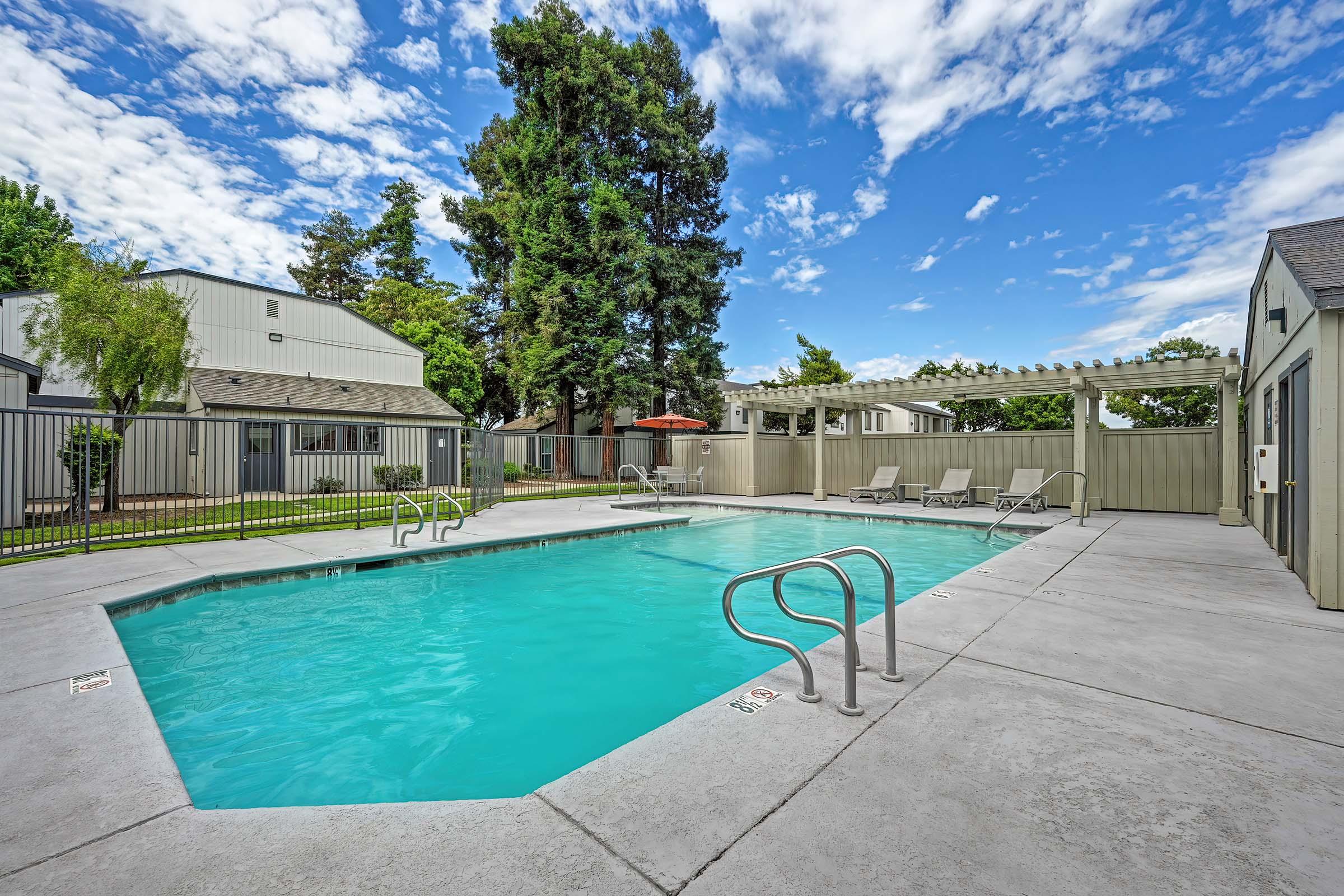 A serene outdoor swimming pool surrounded by trees and a fence. Sun loungers line the poolside, and a shaded area with a pergola provides a relaxing space. The clear blue sky and fluffy clouds add to the inviting atmosphere of the pool area.