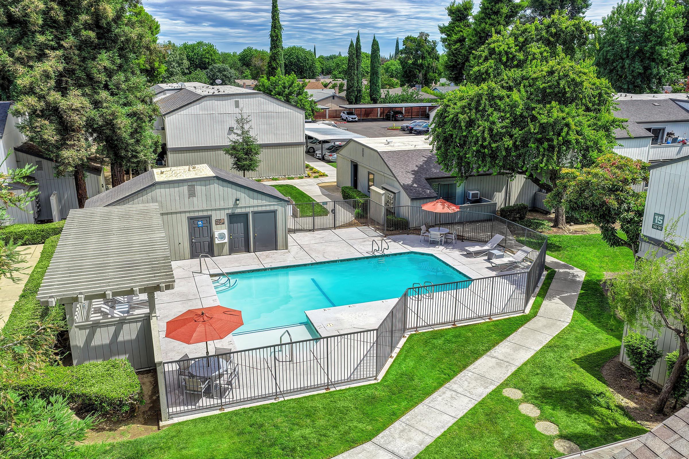 Aerial view of a residential community featuring a swimming pool surrounded by a fence. Sun loungers and two red umbrellas are positioned by the poolside. Lush greenery lines the area, with several buildings in the background and a parking lot visible. The sky is partly cloudy, creating a bright and inviting atmosphere.
