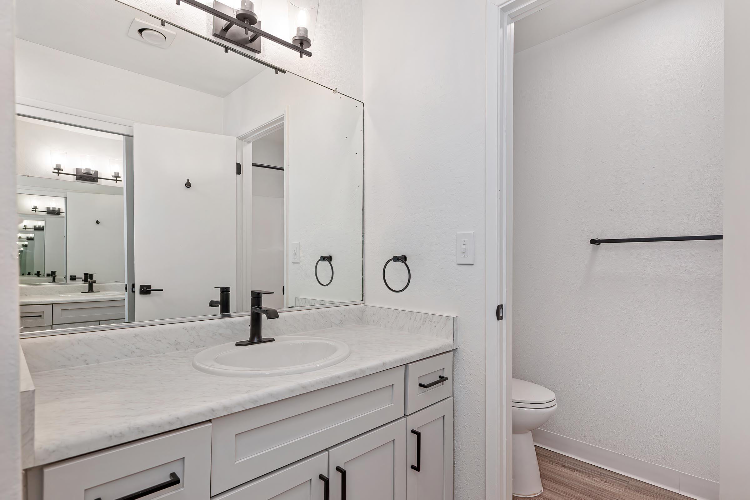 A modern bathroom featuring a marble countertop sink with a black faucet, large mirror, and overhead lighting. The space includes white cabinetry, a towel ring, and a separate small room with a toilet. The walls are painted white, and the flooring is light-colored wood.