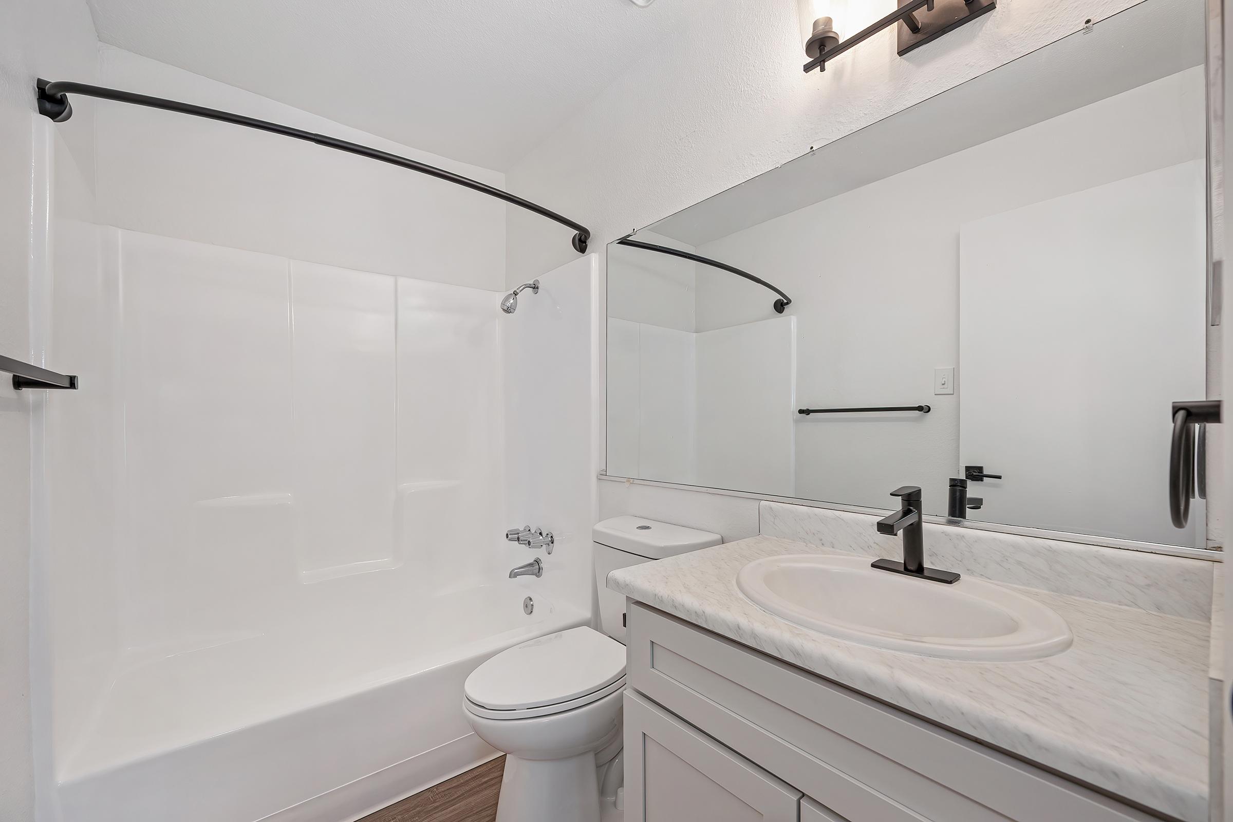A modern bathroom featuring a clean white bathtub and shower combination, a sleek vanity with a marble countertop, a circular sink, and a mirror. The walls are painted in a light color, and there is a black towel rack and faucet for a contemporary touch. Wood-look flooring completes the aesthetic.