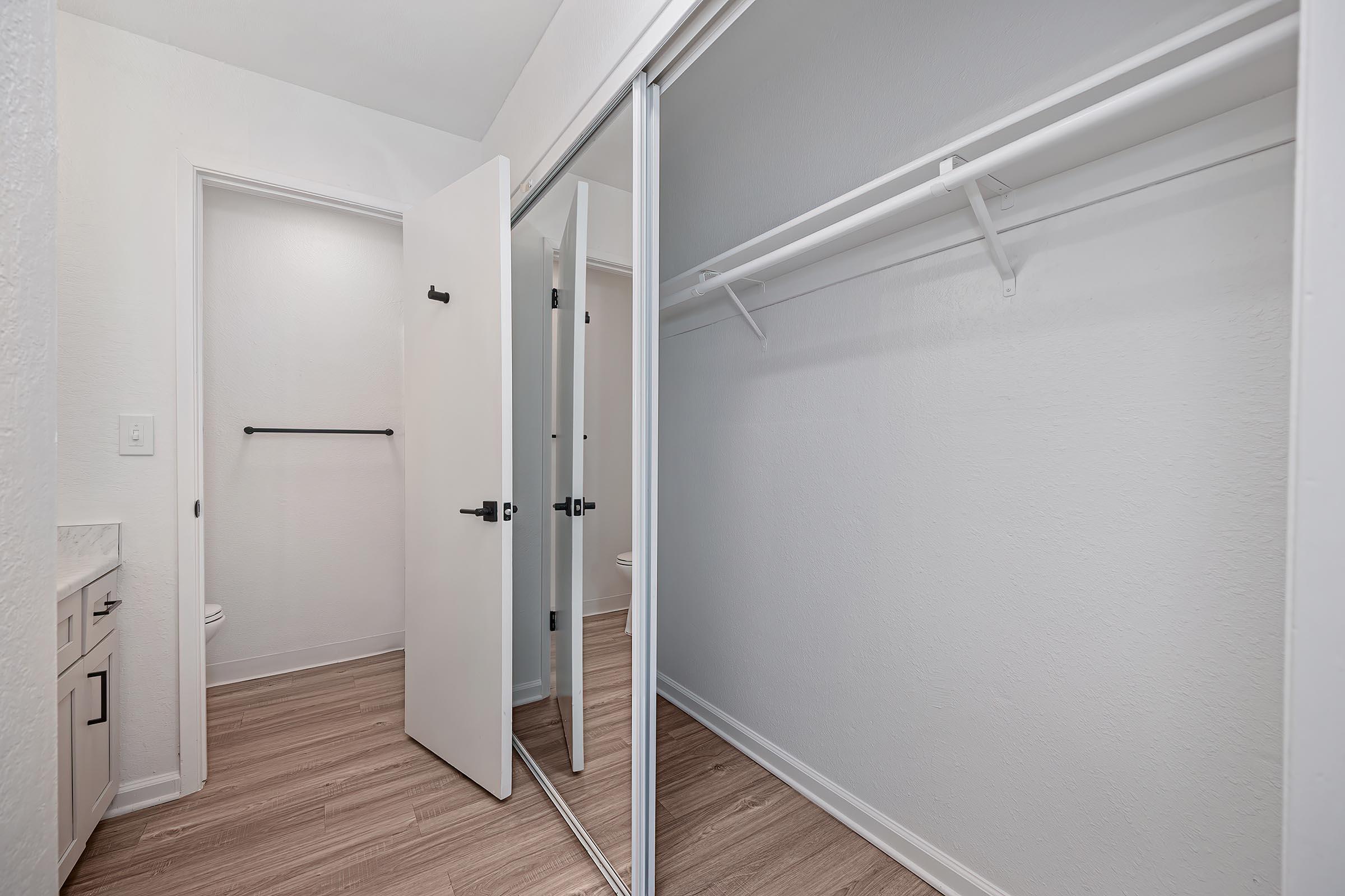 Interior view of a closet featuring mirrored sliding doors, a hanging rod, and a light-colored wooden floor. The space is bright with white walls and includes a partially visible bathroom area to the left.