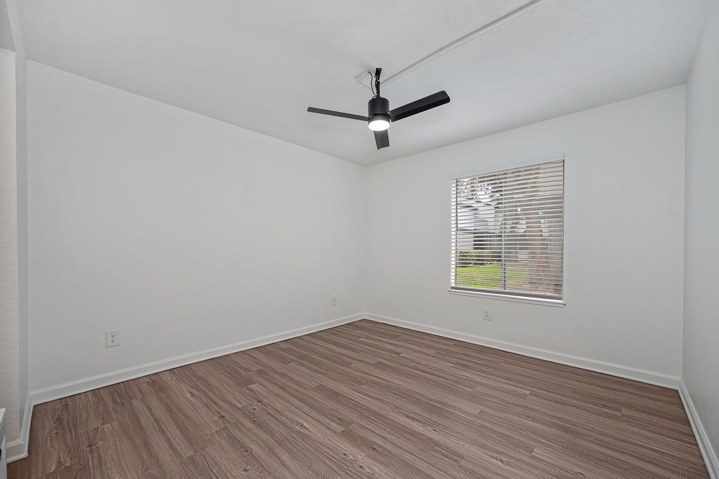 Empty room featuring light-colored walls, a ceiling fan with a light fixture, and a window with blinds. The floor is made of wood-like laminate, and the overall space is bright and airy, with natural light coming from the window.