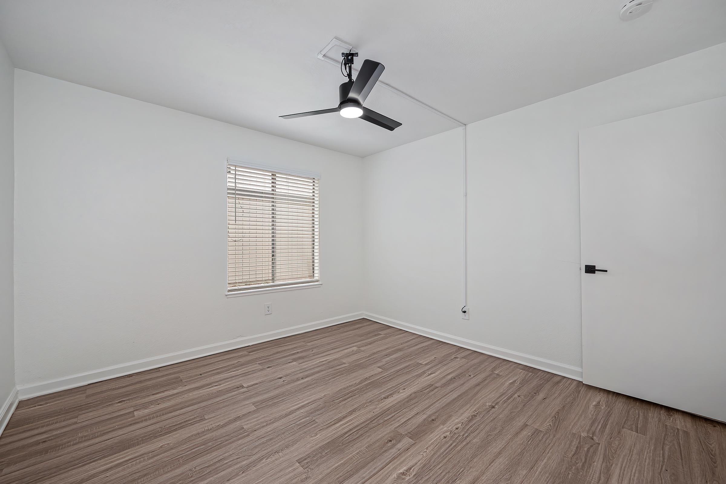 A well-lit empty room with a light-colored wall and wooden flooring. It features a ceiling fan and a window with blinds, allowing natural light to enter. The room includes a plain door and a minimalist design, creating a spacious and clean atmosphere.