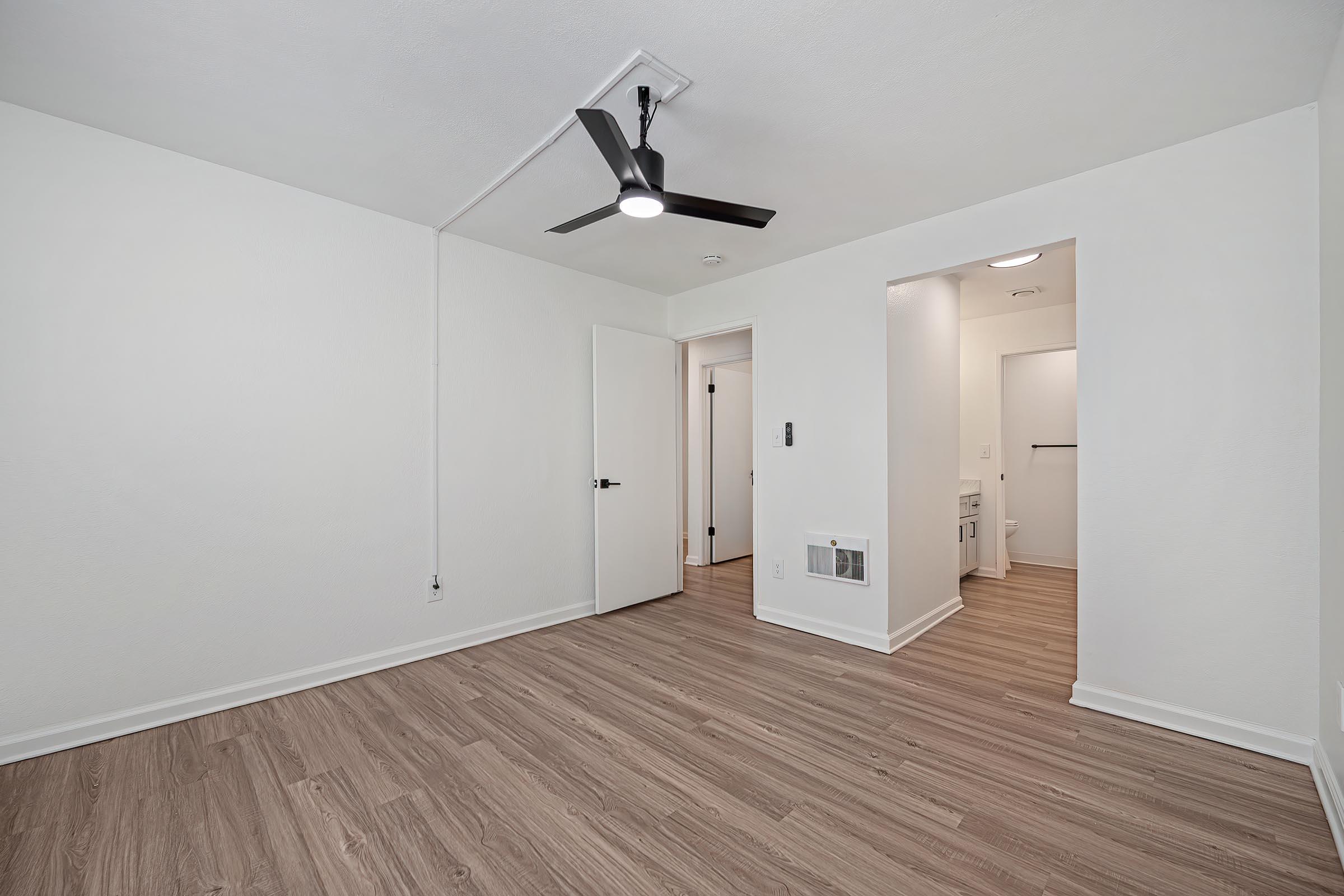 Empty room with light-colored walls and light wood flooring. A modern ceiling fan is installed, and there are two doors visible—one leading to a bathroom and another to an adjoining space. The room is well-lit and appears spacious, creating a clean and inviting atmosphere.
