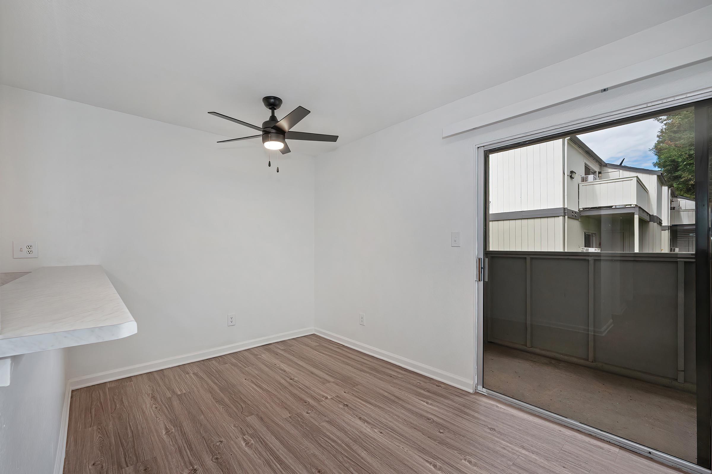 Interior view of a room featuring a ceiling fan, light-colored walls, and a sliding glass door leading to a balcony. The floor is wooden laminate. The space appears bright and empty, suitable for furniture arrangement.