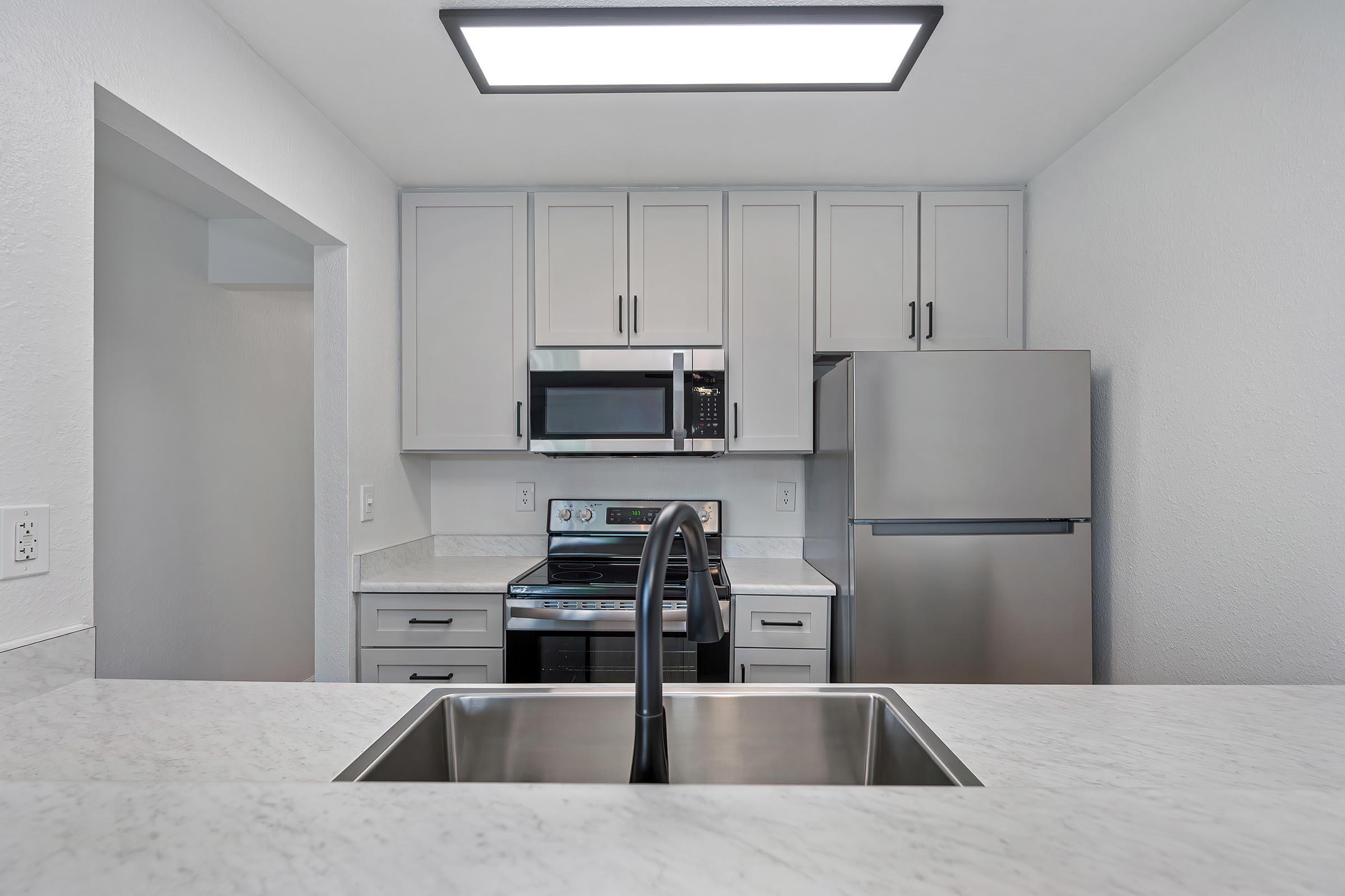 Modern kitchen featuring white cabinetry, stainless steel appliances including a microwave and refrigerator, and a stainless steel sink with a modern faucet. The space has marble countertops and is well-lit by overhead lighting.