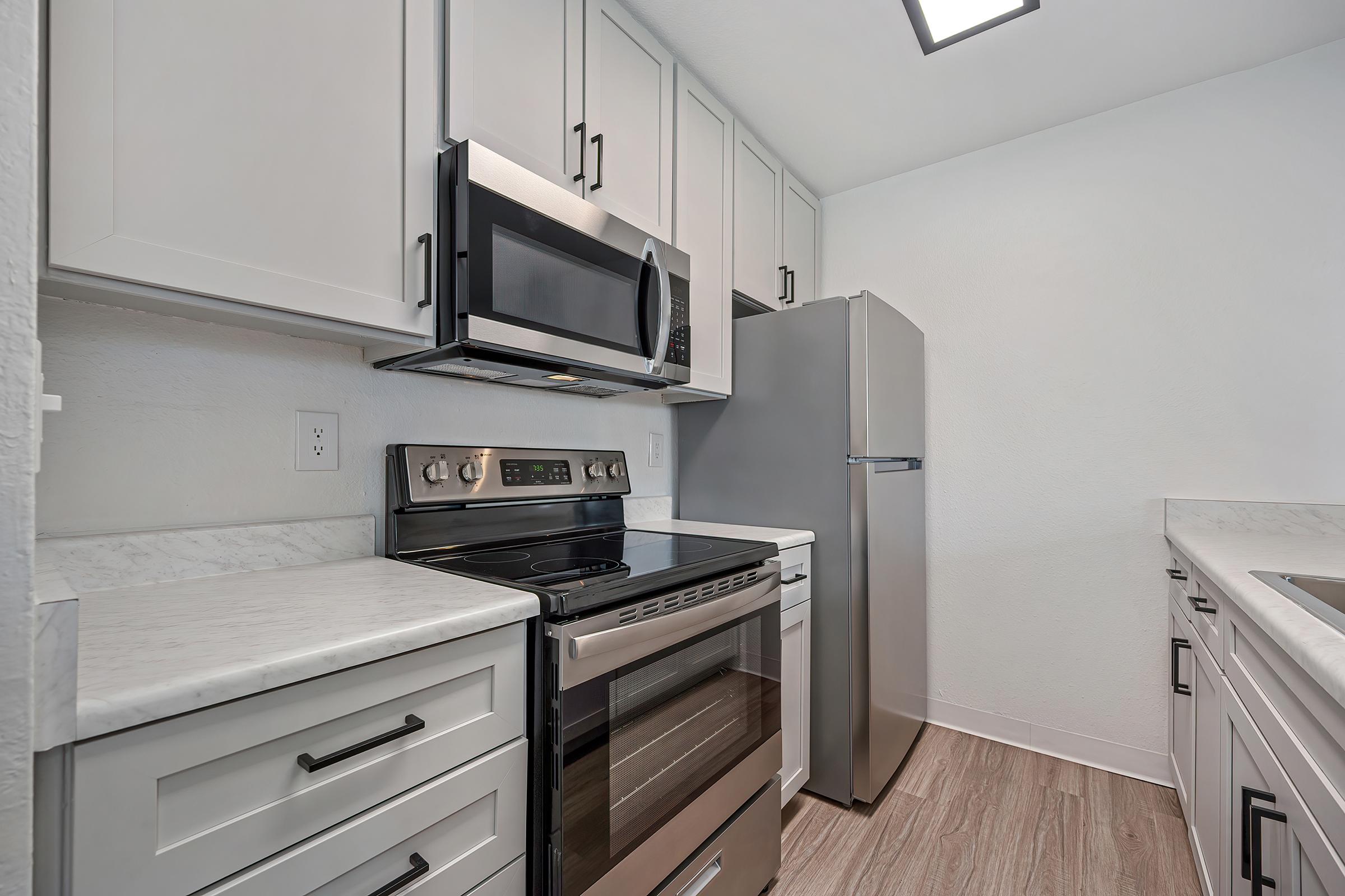 Modern kitchen featuring stainless steel appliances, including an oven, microwave, and refrigerator. White cabinets with sleek hardware and a marble-like countertop. The walls are painted in a light color, and the flooring has a wood-like design, creating a clean and contemporary look.