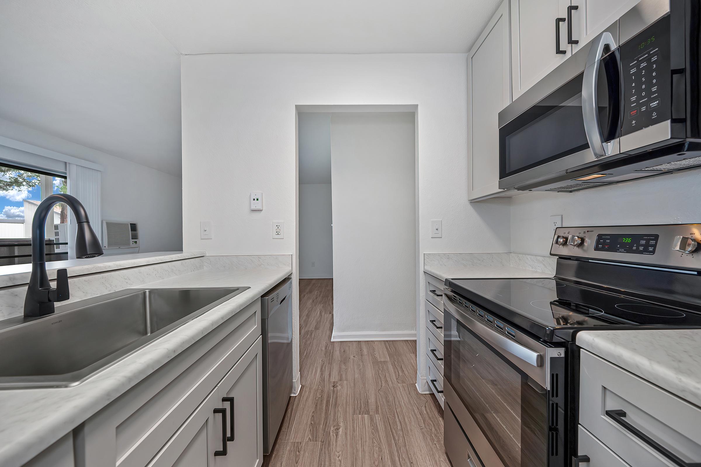 Modern kitchen featuring stainless steel appliances, including a microwave and stove. The countertops are light-colored with a sink and faucet. The space has a bright ambiance with white walls and light wood flooring, leading to an open hallway in the background. Natural light illuminates the area.