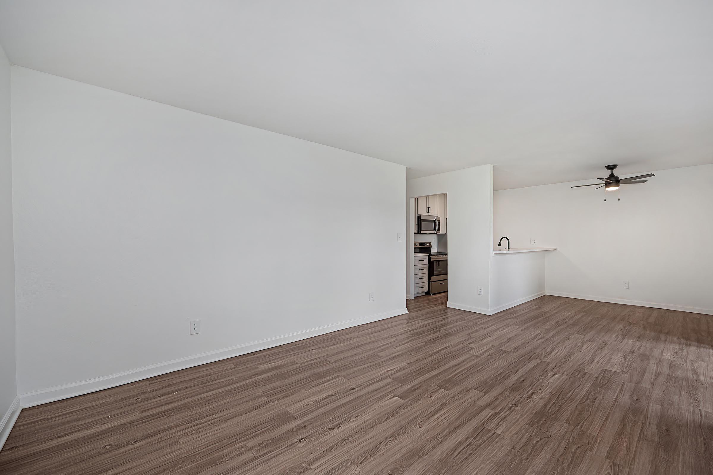 A spacious, empty room with light-colored walls and wooden flooring. It features a ceiling fan and an open layout that connects to a kitchen area in the background, showcasing modern appliances and cabinetry. The overall ambiance is bright and inviting, ideal for new tenants to personalize.