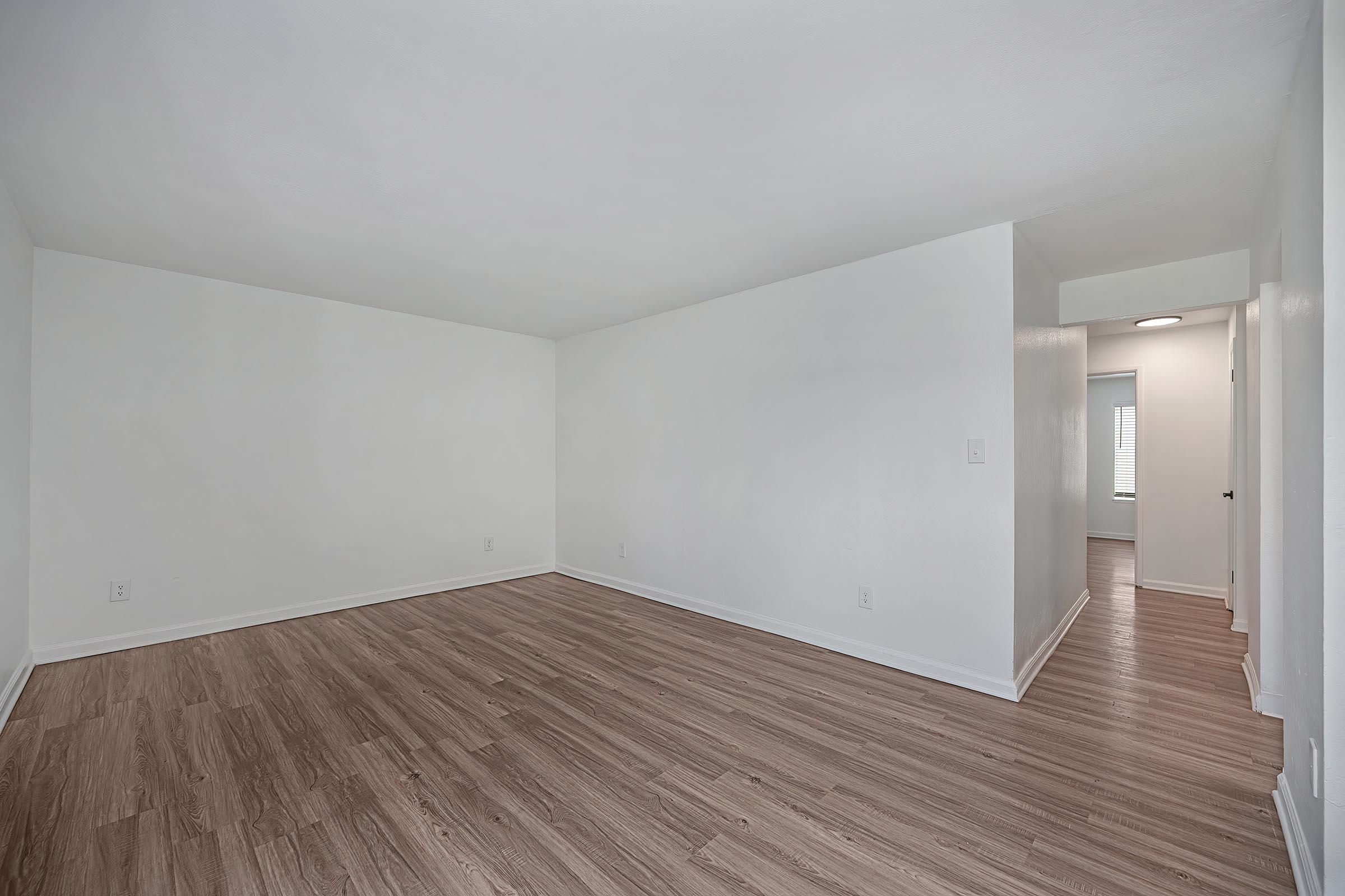 A spacious, empty room with white walls and light brown laminate flooring. Natural light streams in from a doorway at the far end, leading to another area. The room has no furniture, emphasizing its openness and clean, minimalist design.