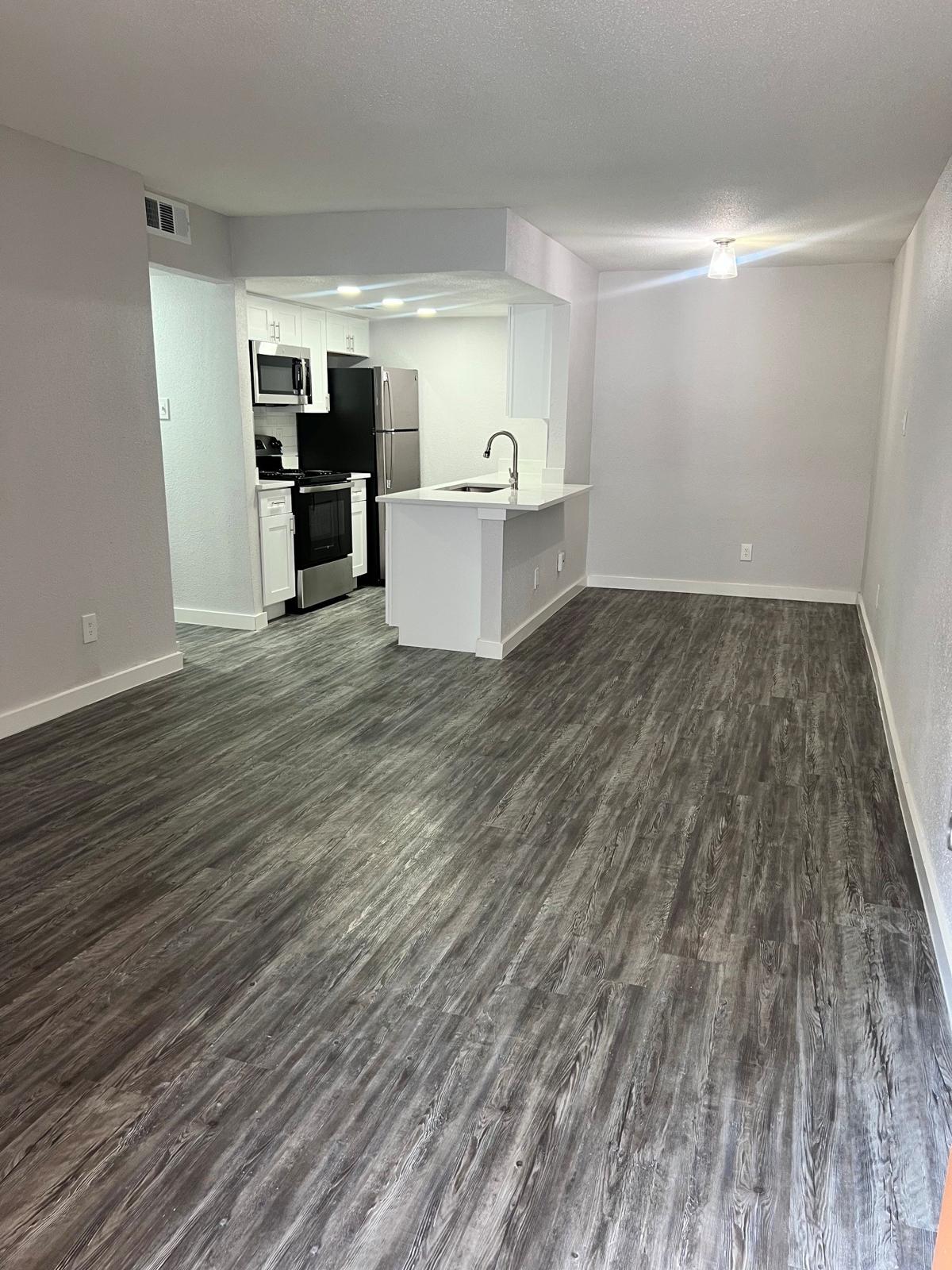 A spacious, modern living area with light-colored walls and sleek, dark wood-like flooring. The open layout features a kitchen with stainless steel appliances, including a refrigerator and oven, and a white countertop. Natural light illuminates the room from a ceiling light fixture.