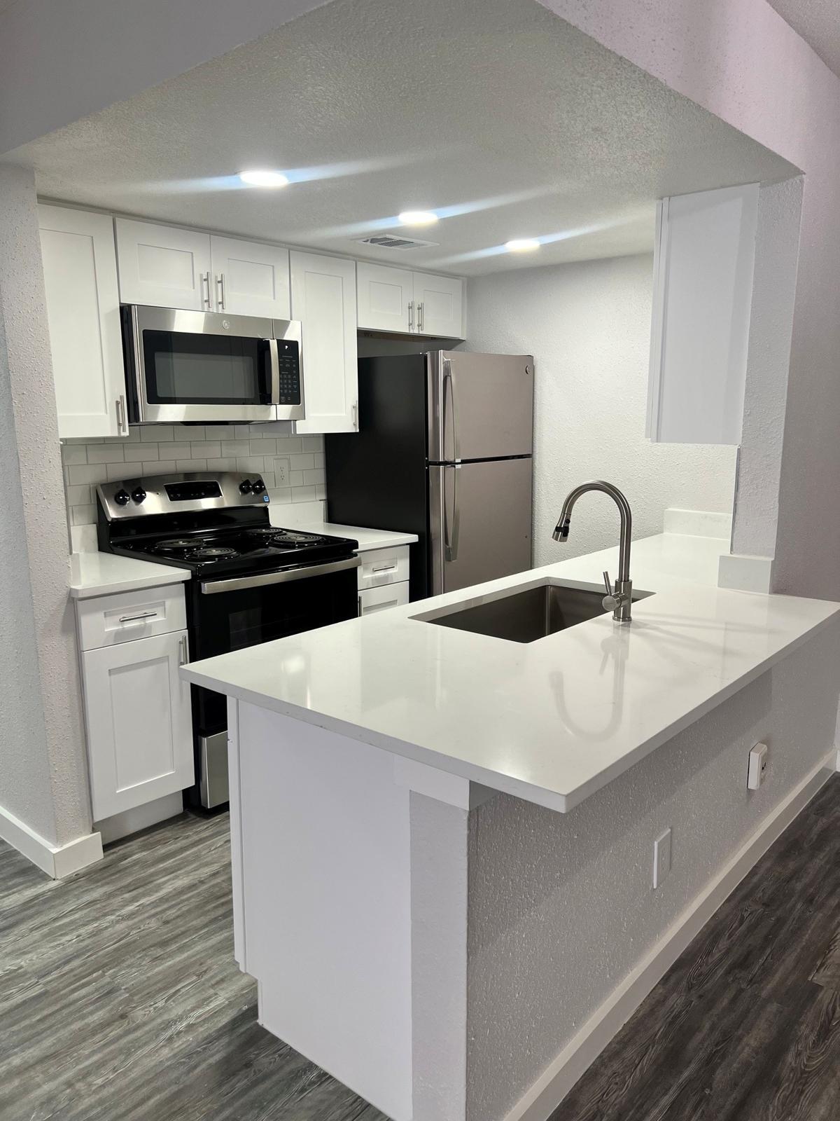 Modern kitchen with white cabinetry, stainless steel appliances including a microwave, stove, and refrigerator. The kitchen features a white countertop and a sink, with an open layout connecting to an adjacent living area. Bright overhead lighting enhances the space.