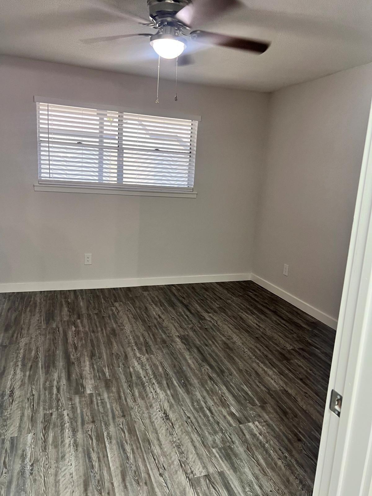 A sparsely furnished room with light gray walls and wood-like flooring. The space features a ceiling fan and a window with white blinds, allowing natural light to enter. The room appears clean and freshly painted, suitable for various uses.