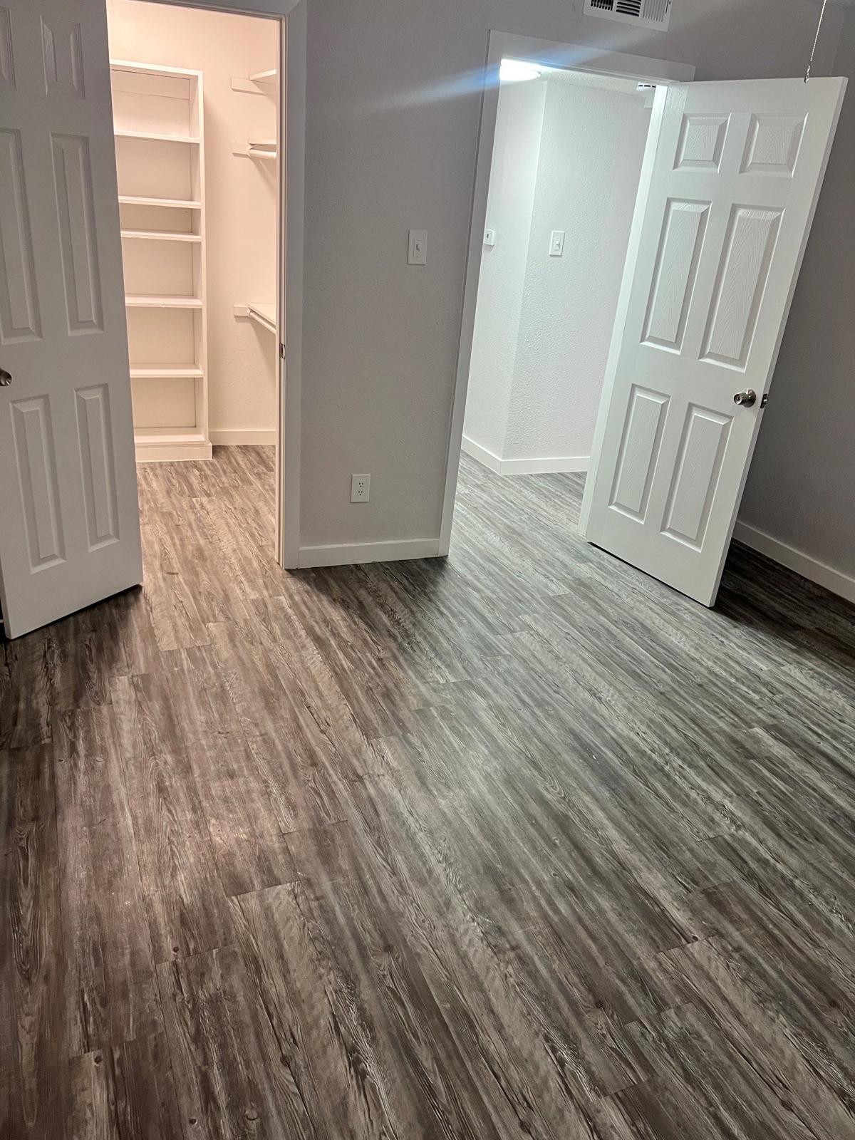 A spacious room with new gray laminate flooring, featuring a doorway leading to a well-lit closet with shelves. Two white doors are visible, and the walls are painted in a light color, enhancing the brightness of the space. Natural light is coming in from the right side.