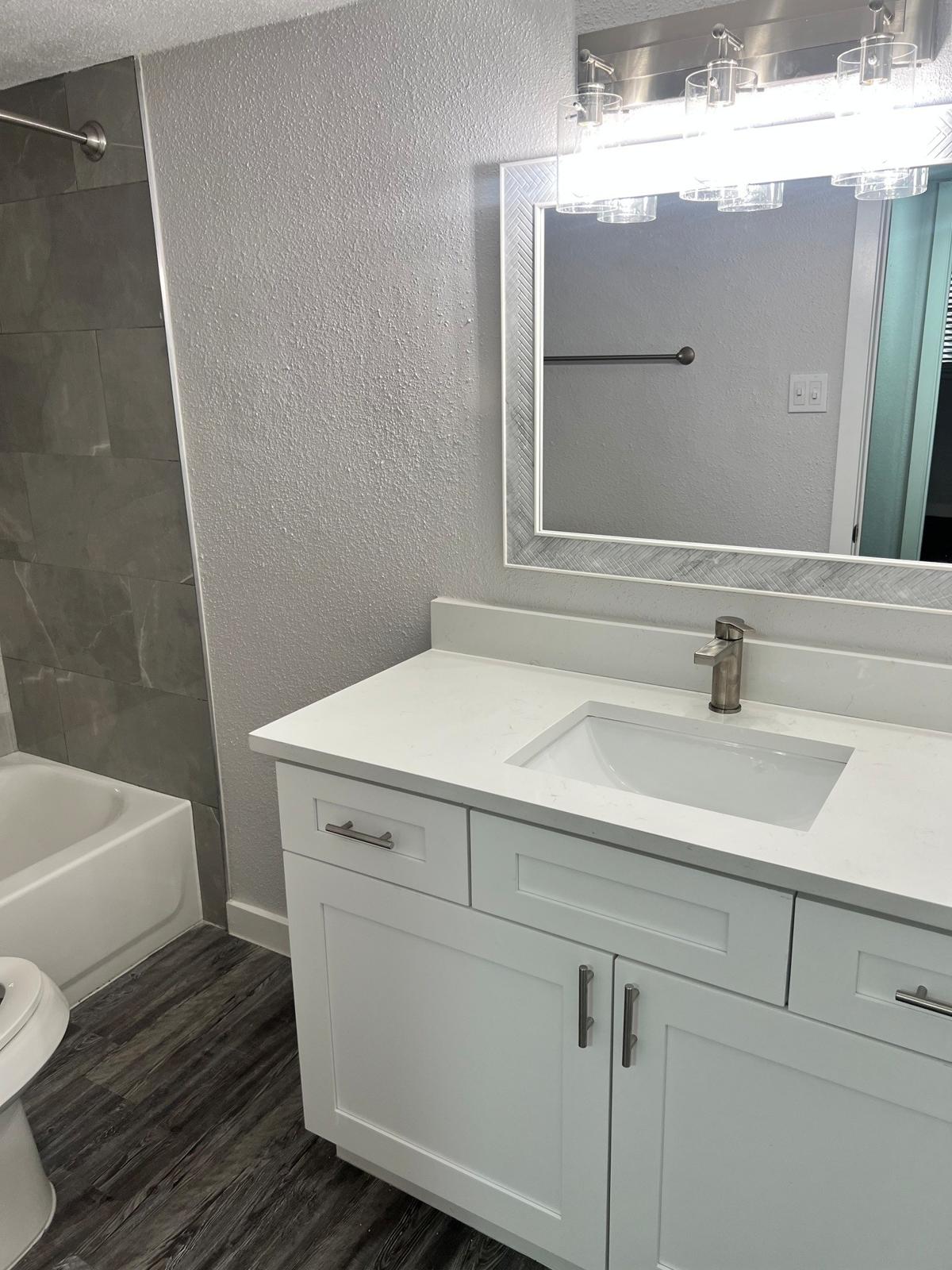 A modern bathroom featuring a white vanity with a sink, a large mirror, and contemporary light fixtures. The space includes a bathtub with a shower, gray tiled walls, and a dark wood floor. A towel bar is mounted on one wall, and bright lighting enhances the fresh and clean atmosphere.