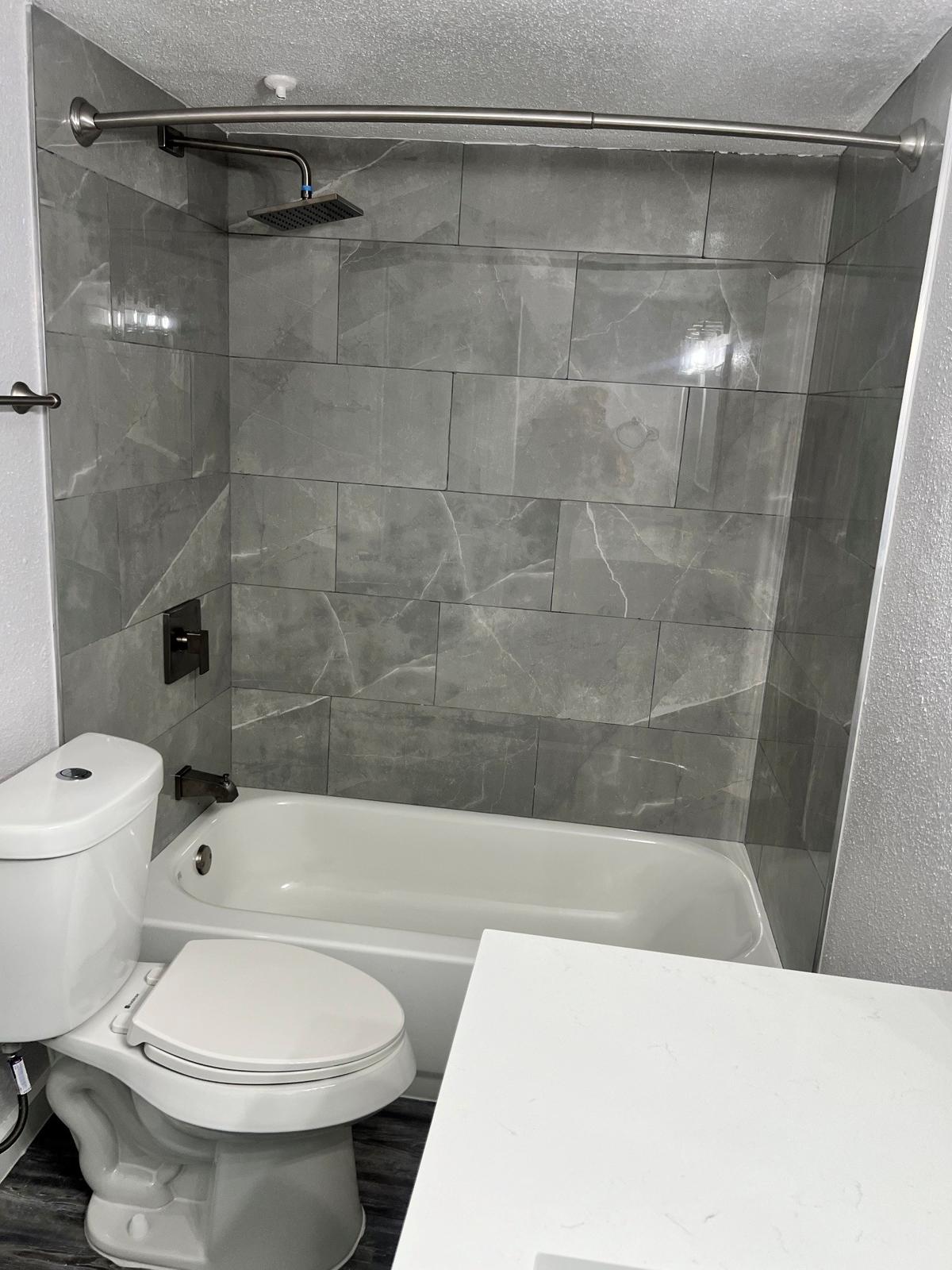 A modern bathroom featuring a gray tiled shower area with a rainfall showerhead, a white bathtub, and a white toilet. The walls are tiled with large gray tiles, and there is a minimalistic white countertop visible in the foreground. The space has a clean and contemporary design.
