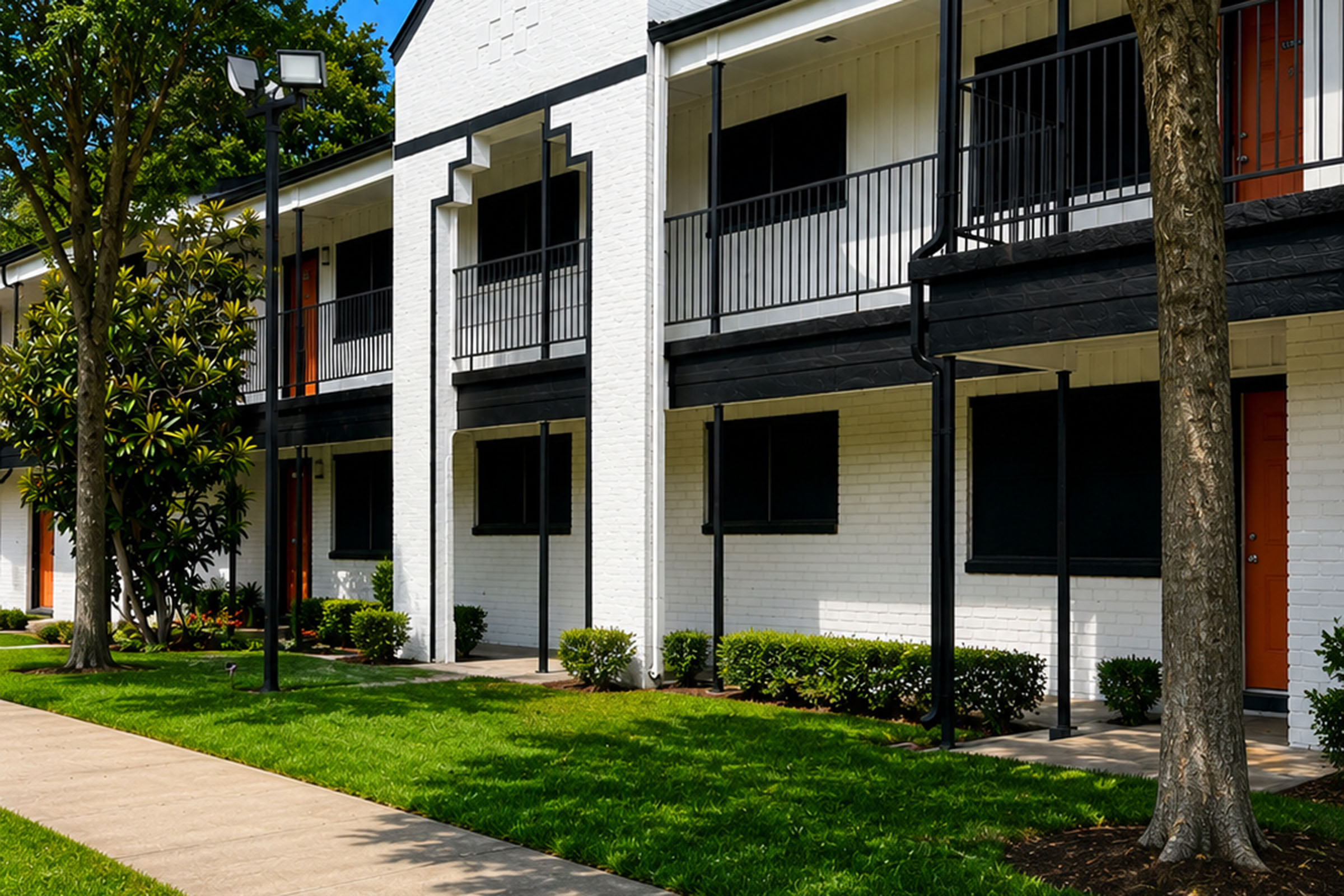A row of white apartment buildings with black railings, surrounded by well-maintained lawns and shrubs. Pathways lead to the entrances, and trees provide shade. The buildings have large windows, but the windows are dark, suggesting they may be unoccupied or have their blinds drawn.