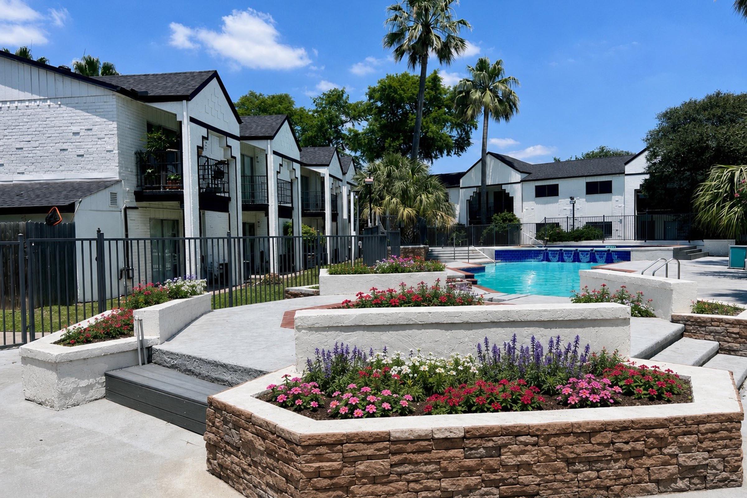 A sunny outdoor area featuring a swimming pool surrounded by landscaped gardens with colorful flowers. The scene includes several apartment buildings in the background, palm trees, and a well-maintained lawn, creating a welcoming and relaxing atmosphere.