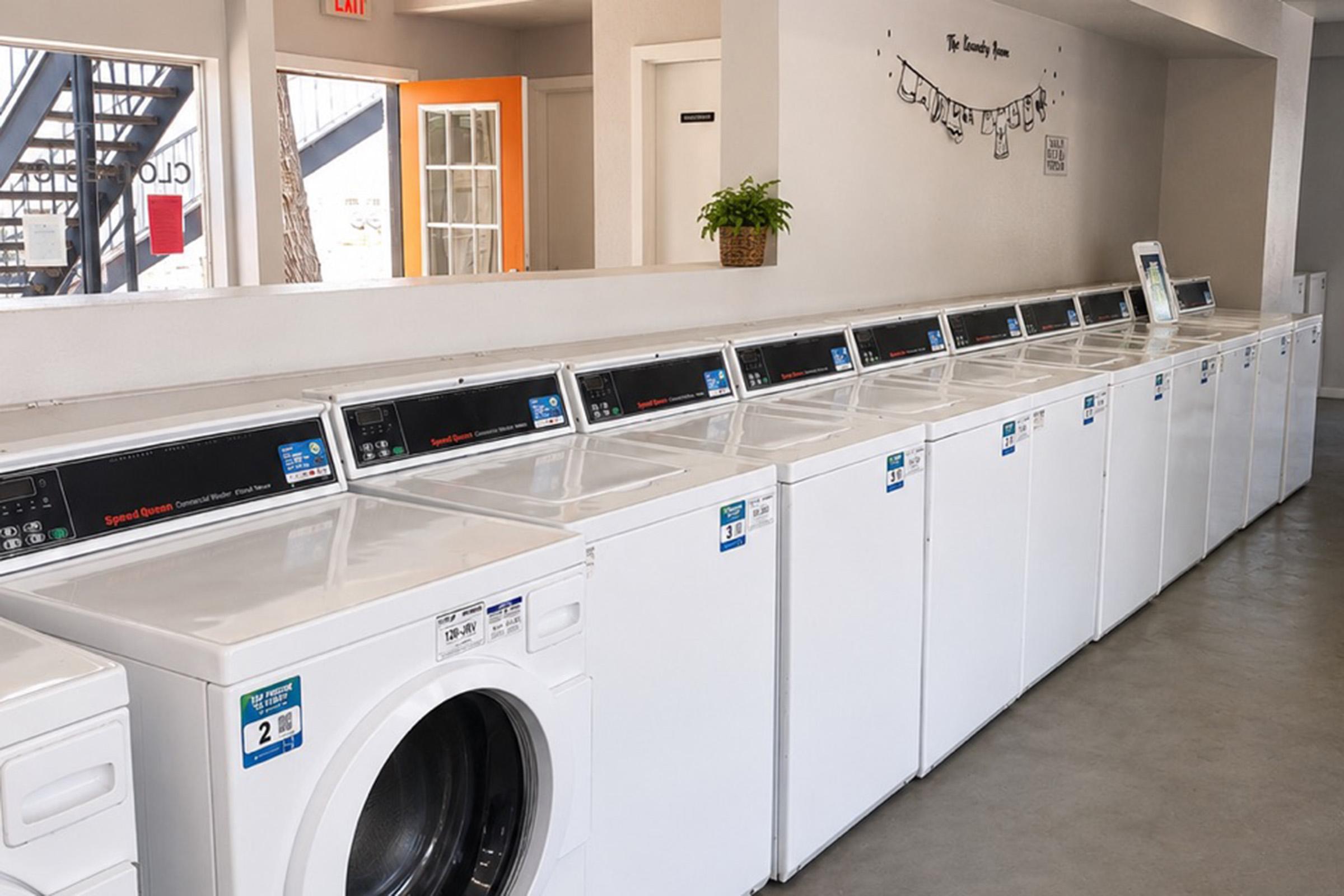 A row of white commercial washing machines and dryers in a clean laundry facility. The machines have digital displays and multiple settings. The interior is bright with a modern design, featuring a potted plant and a decorative sign on the wall. Clear pathways for easy access between machines.