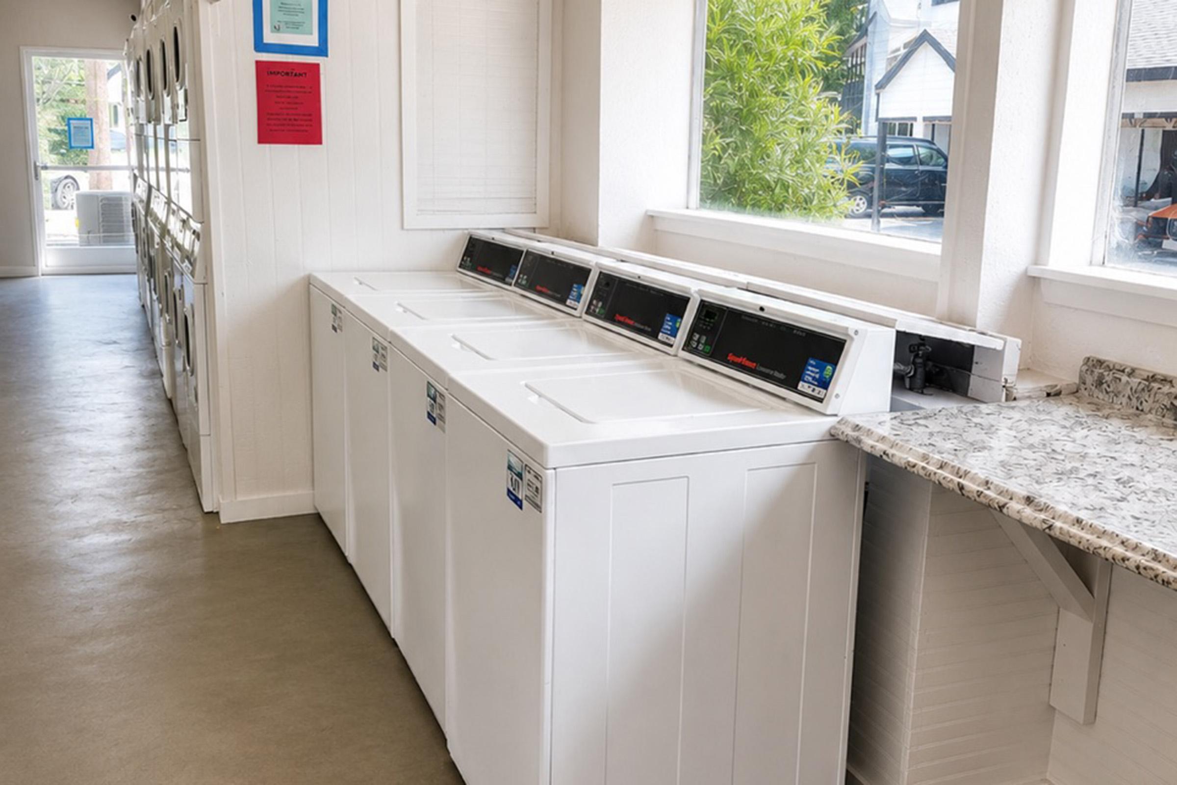 A row of white washing machines in a bright laundry room, featuring large windows that let in natural light. The machines are lined up against the wall, and a countertop made of textured stone is visible beside them. Informational signs are displayed on the walls, enhancing the functional space.