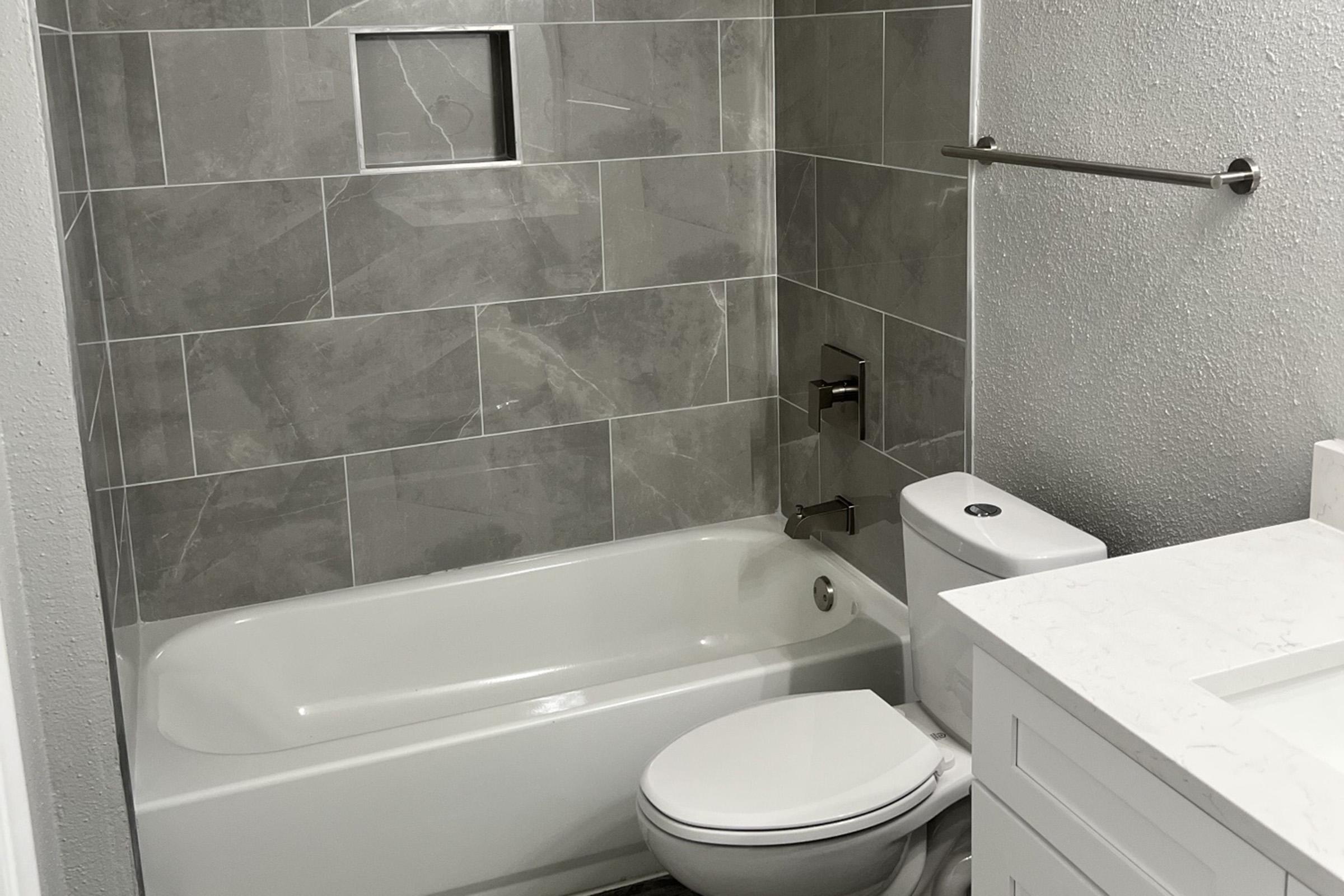 A modern bathroom featuring grey tiled walls, a white bathtub with a shower, and a toilet. There's a small recessed shelf in the wall and a light-colored countertop with a sink. The overall design is clean and contemporary.
