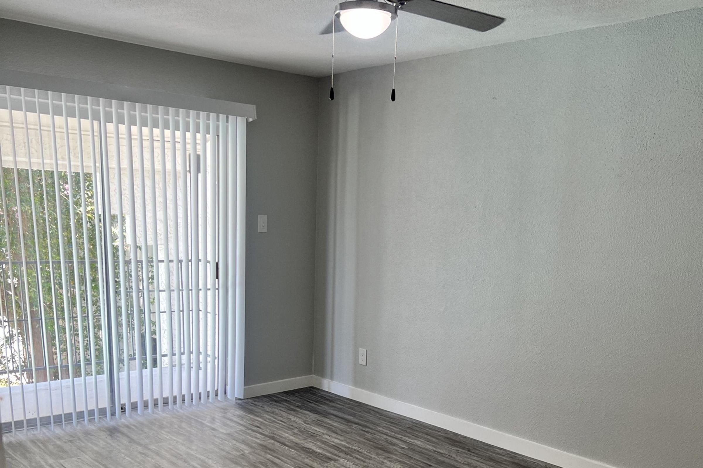 A bright, empty room with light gray walls and a ceiling fan. Large sliding glass doors with vertical blinds allow natural light to enter, leading to an outdoor space. The floor is covered with light wood-like laminate. The room has a clean and modern appearance, perfect for personalization.