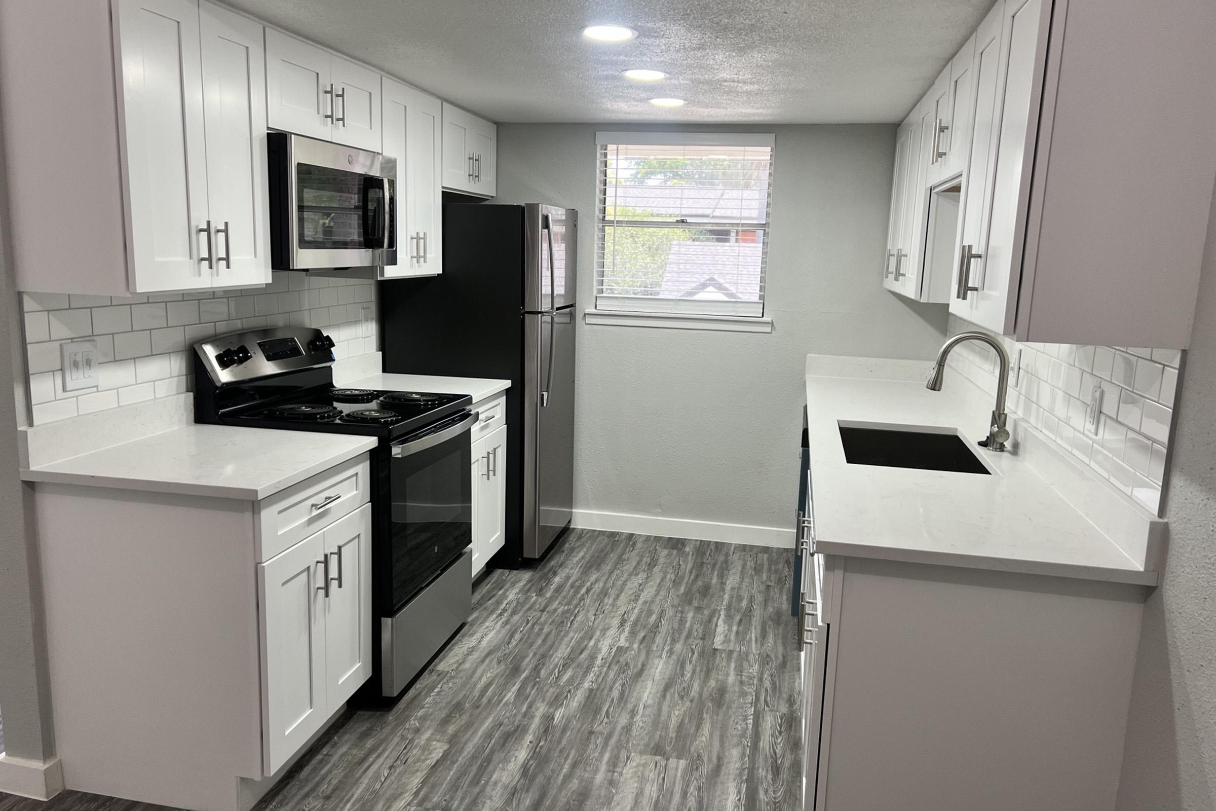 Modern kitchen featuring white cabinets, stainless steel appliances, and a black refrigerator. The countertops are light-colored, and there's a stainless steel sink. Bright lighting and a window enhance the space, which has a wood-like floor. Clean and contemporary design with ample storage.