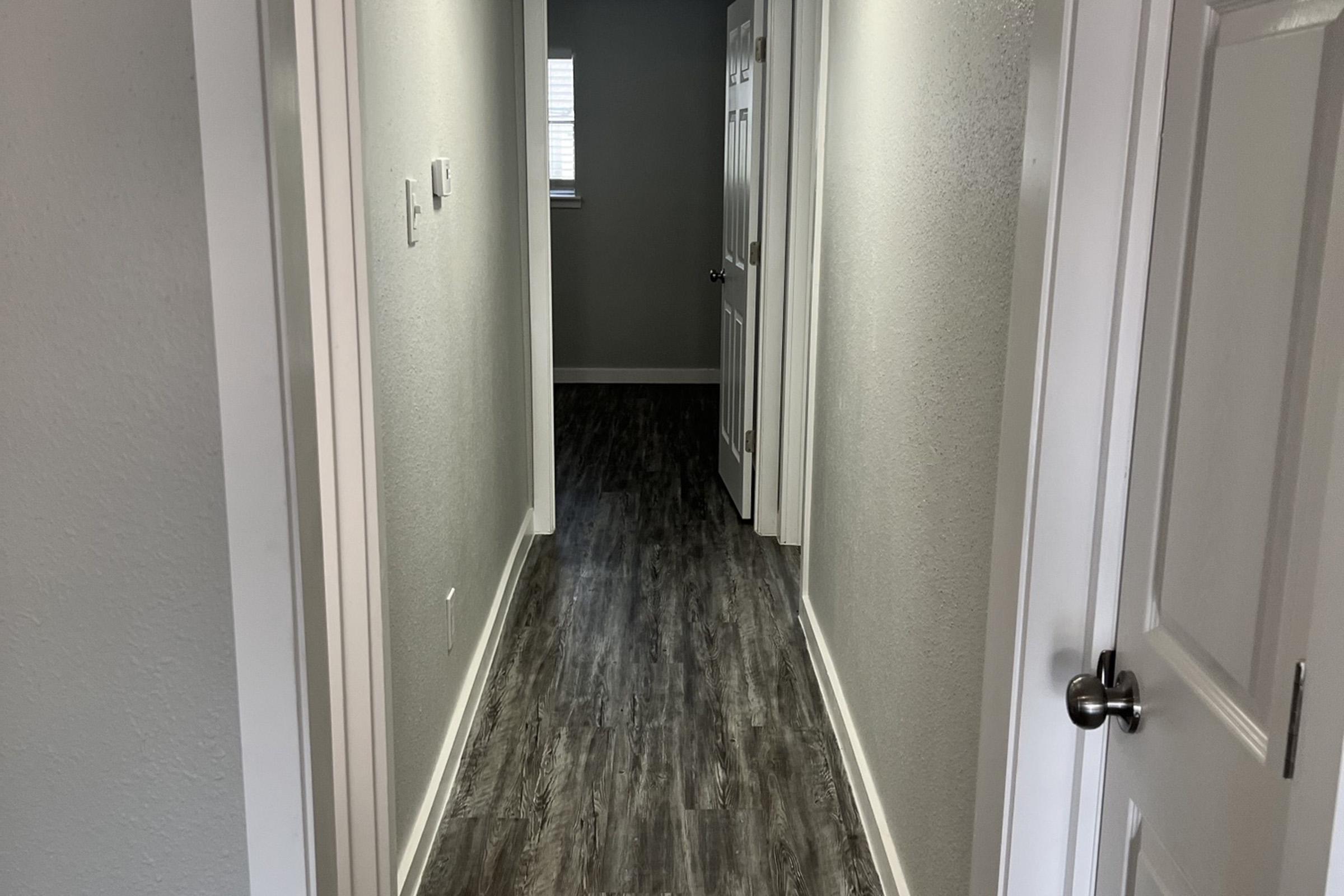 A long, narrow hallway with light gray walls and dark wood-like flooring. There are doors on either side, and a window at the end of the hall allowing natural light to enter. The space is well-lit and appears to be part of a modern interior design.