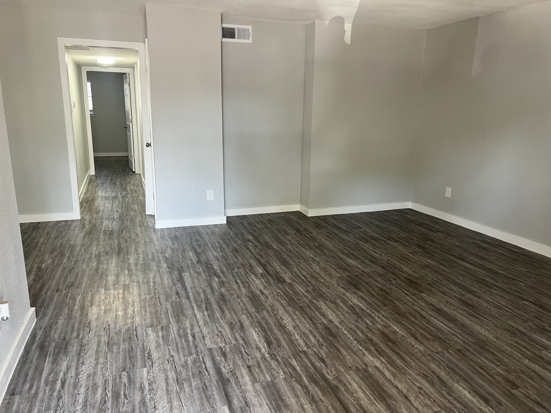 Spacious empty room with gray walls and laminate flooring. A hallway is visible in the background, leading to another room. Natural light enters from a window, highlighting the clean, modern aesthetic of the space. Ideal for customization or as a living area.