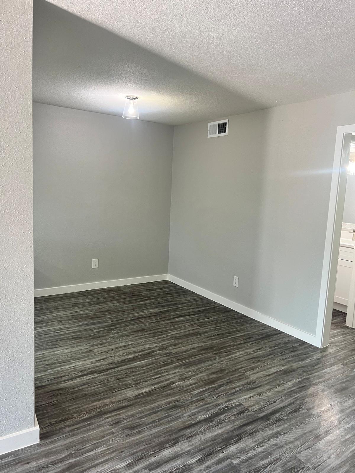 An empty room with light gray walls and a textured wooden floor. A ceiling light is on, providing illumination. The room appears spacious with no furniture, and a doorway leads to a bathroom or another room on the right side.